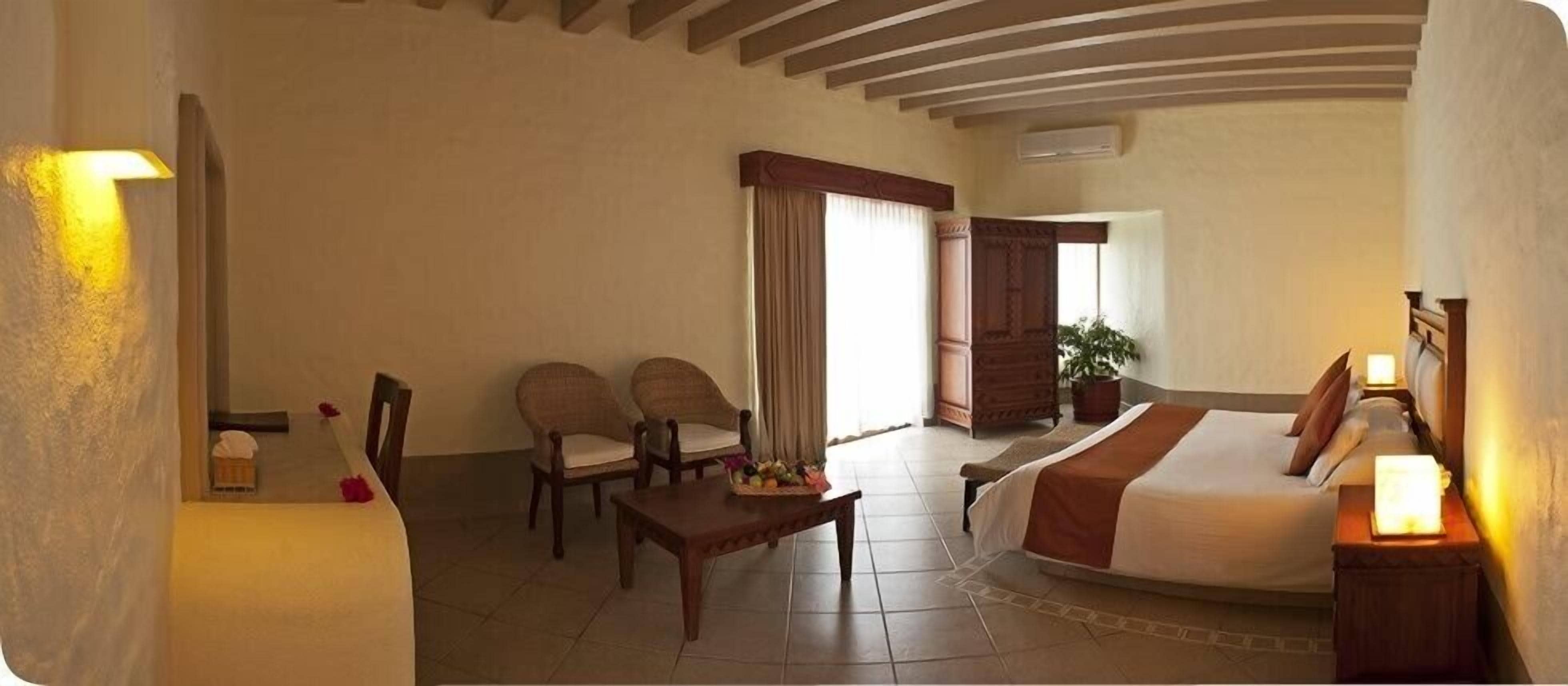 A hotel room with a bed, two chairs, a small table with fruit, a potted plant, and a view of the outside.