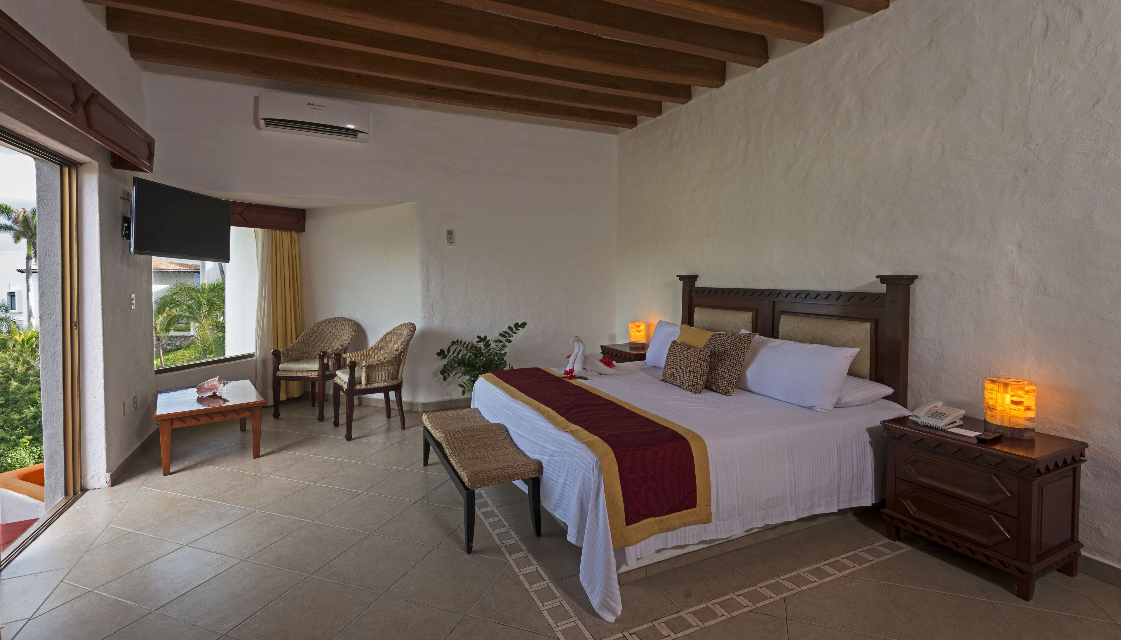 A hotel room with a bed, bedside tables, a chair, a small table, and a window with a view of palm trees.