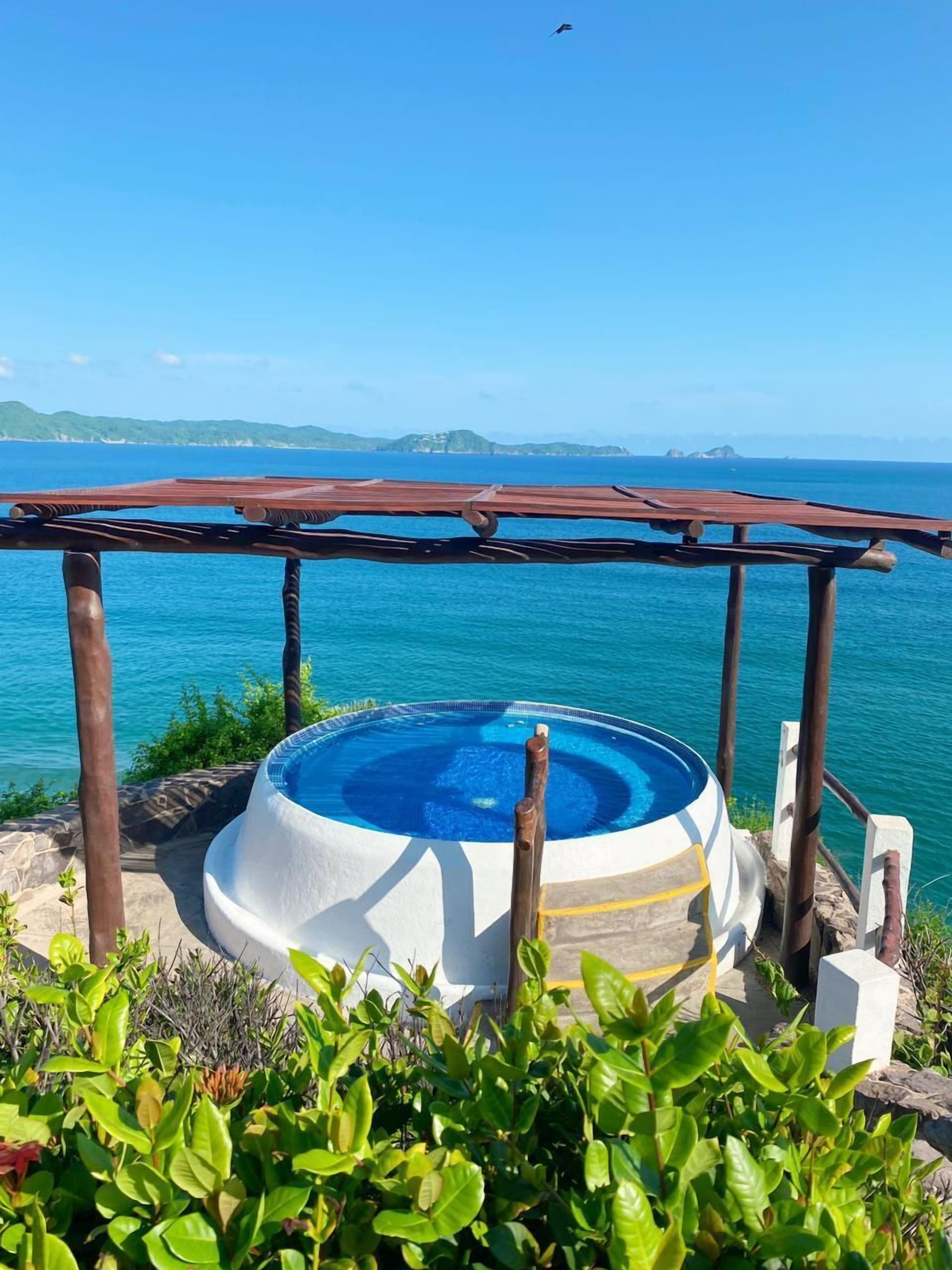 Una vasca idromassaggio con una pergola in legno, immersa nel verde e con vista su un corpo d'acqua e sulla terraferma in lontananza.