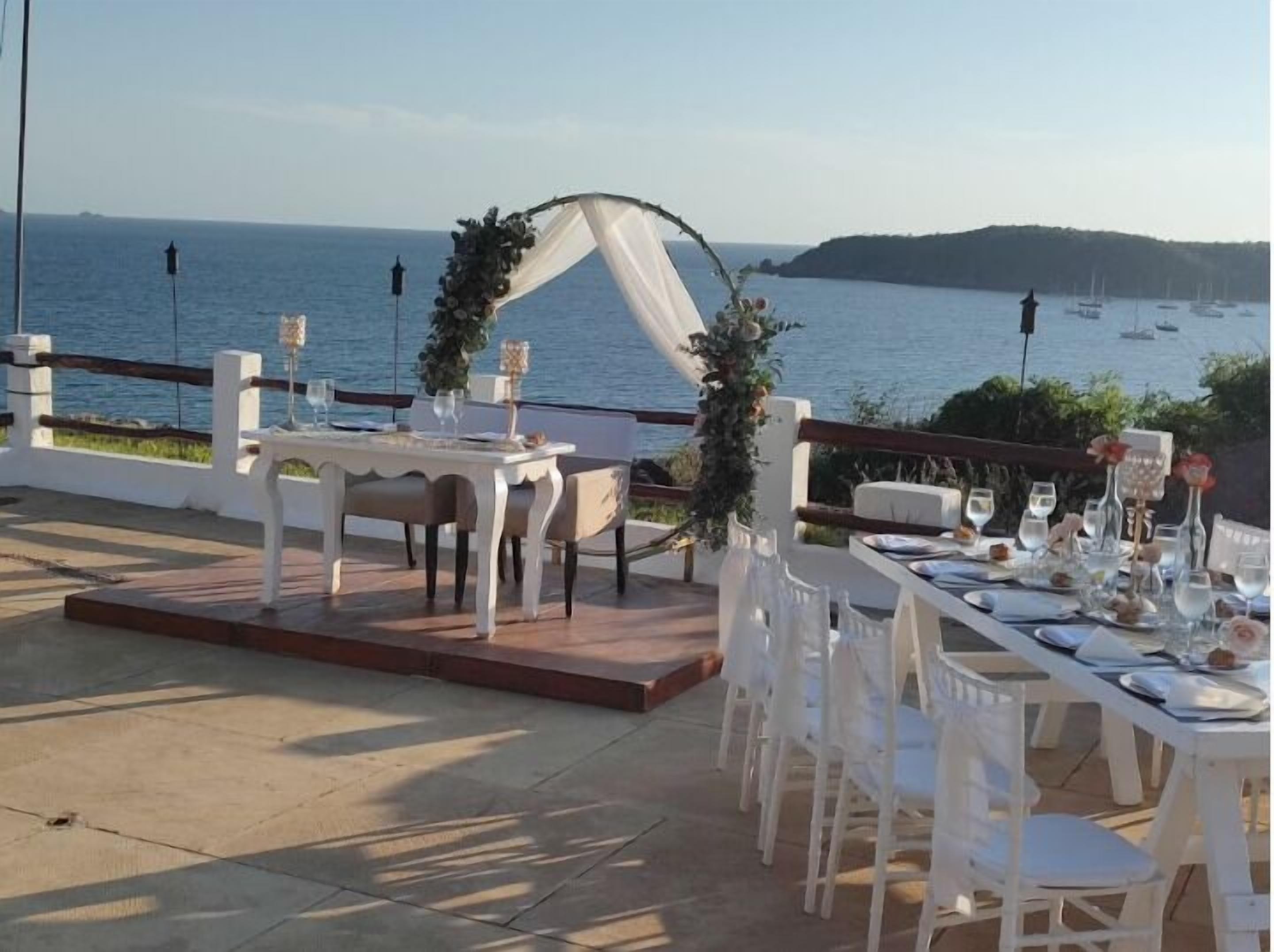 An outdoor dining area with white tables and chairs, a white archway, and a view of the sea.