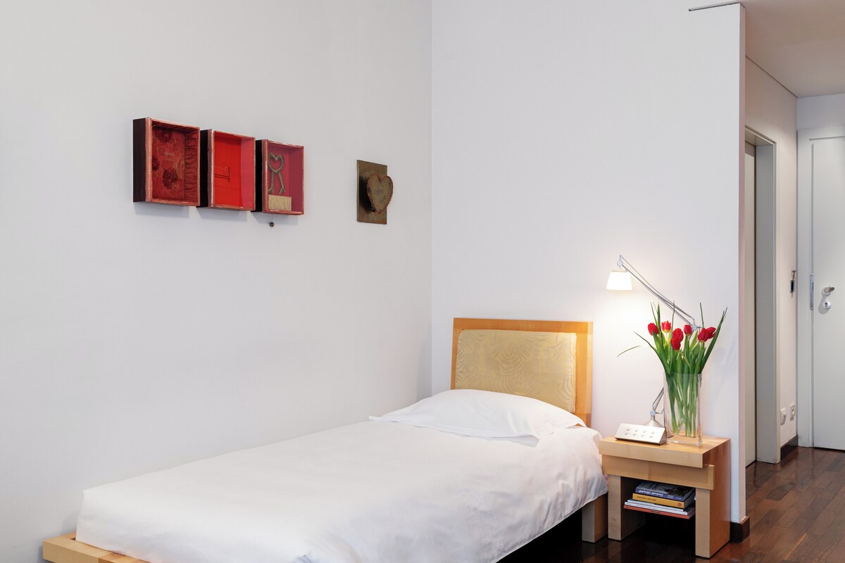 A neatly made bed with a wooden headboard, a bedside table with a lamp and a vase of flowers, and a small shelf with books.