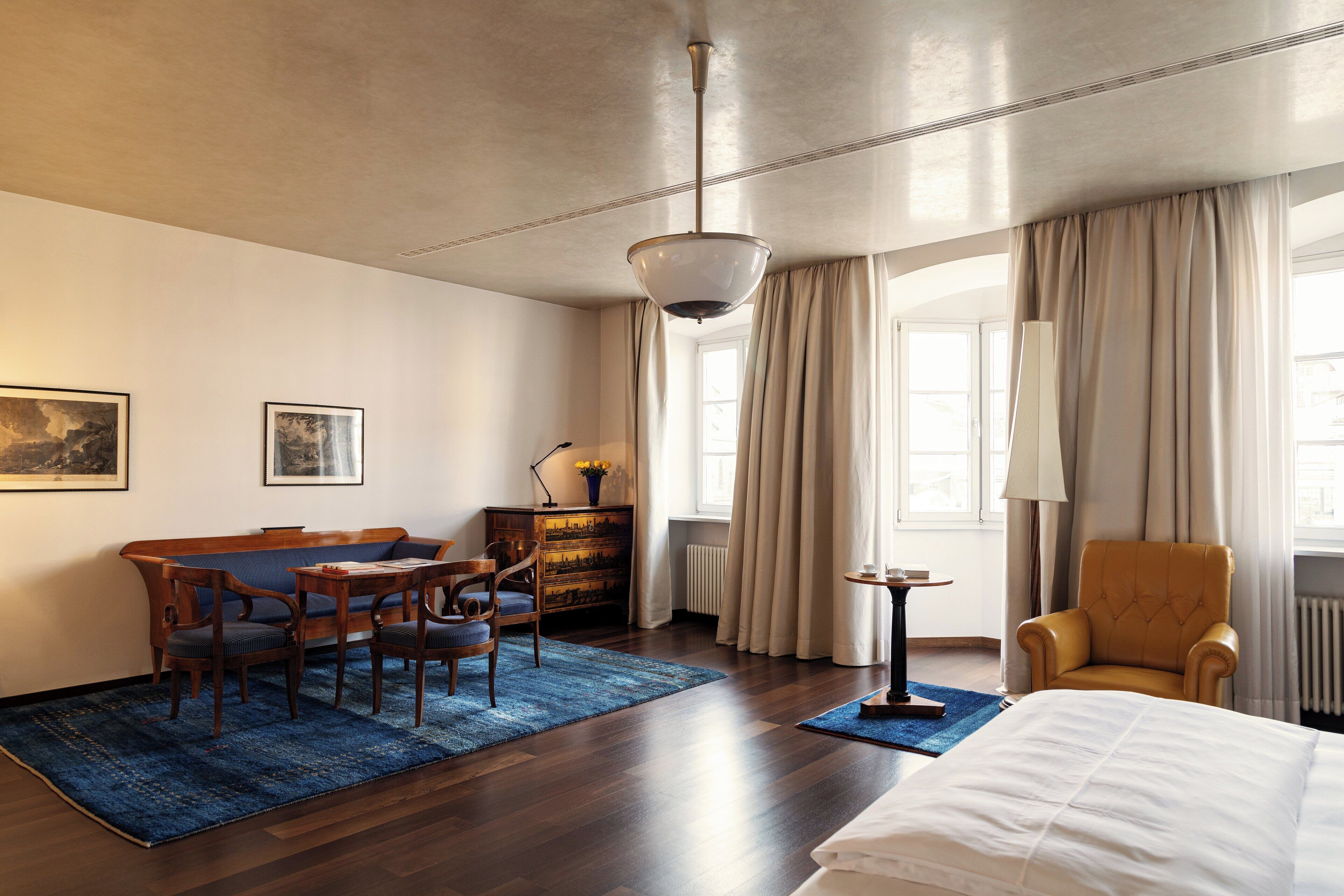 A hotel room with a wooden floor, a bed, a sofa, a desk, and a chair. There is a window with curtains and a lamp on a side table.