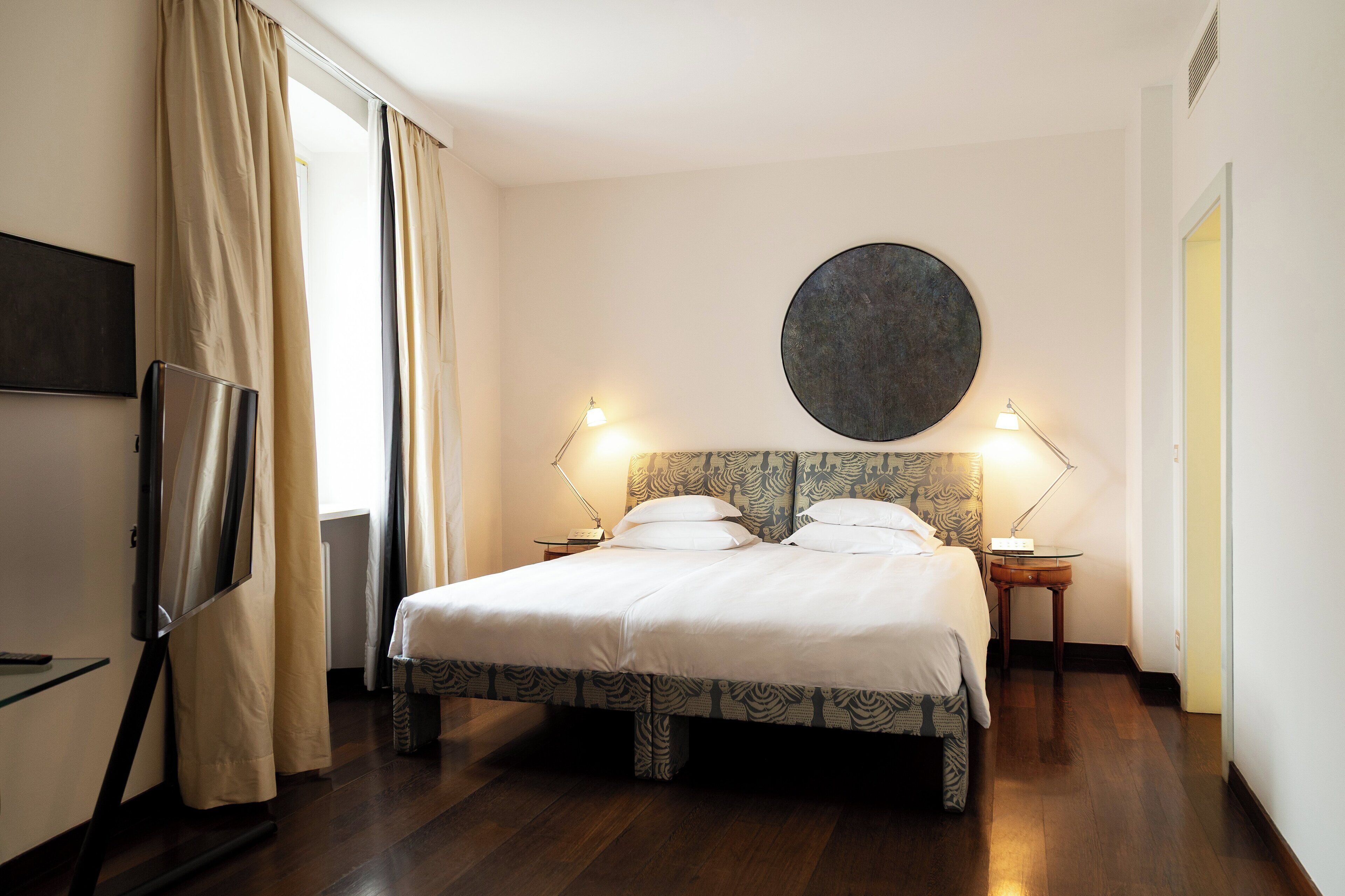 Una cama bien hecha con cabecera, dos mesitas de noche con lámparas, un televisor de pantalla plana en la pared y una obra de arte circular grande sobre la cama.