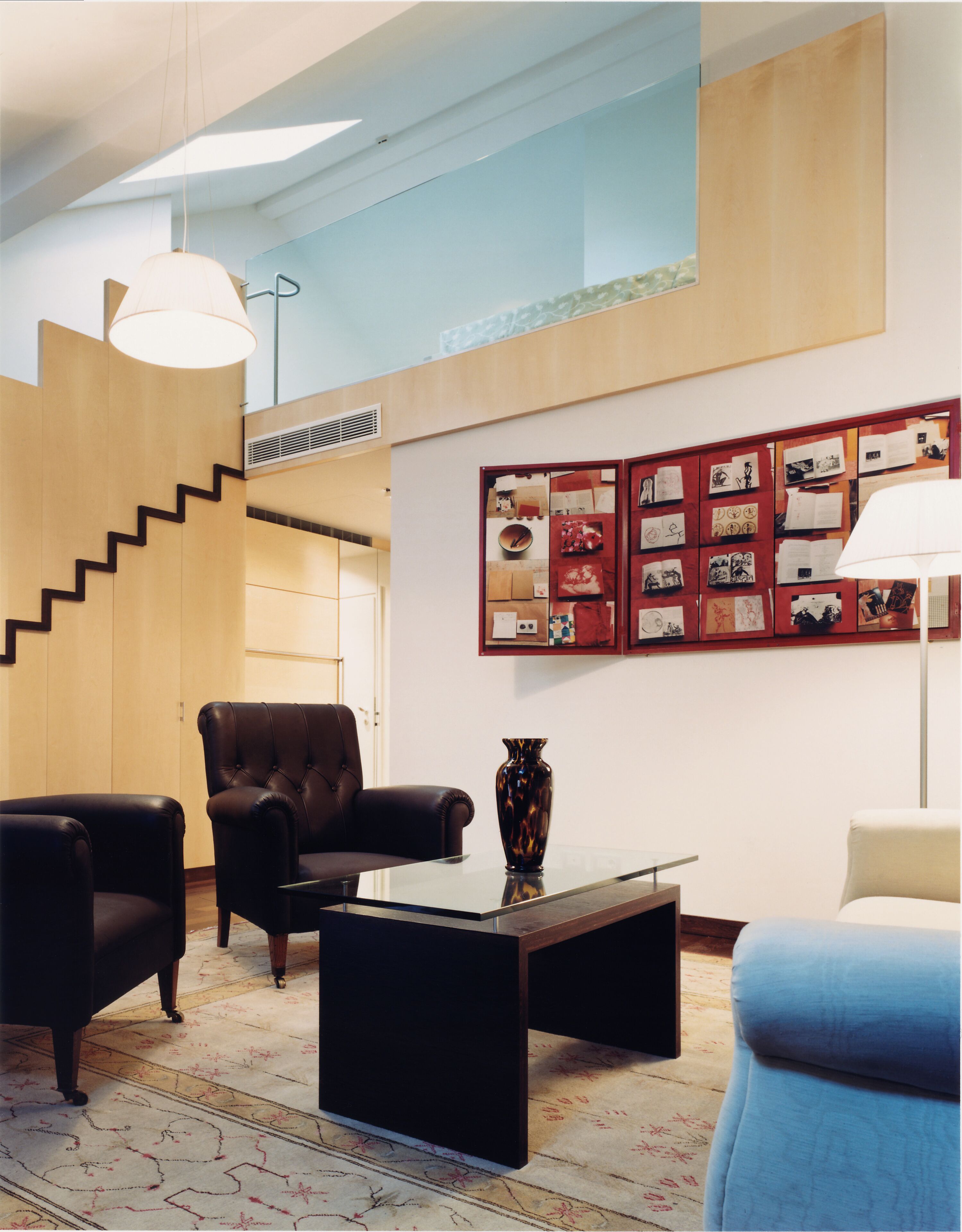 Una sala de estar moderna con una escalera, sillones de cuero, una mesa de centro de vidrio y una pared decorada con cuadros.