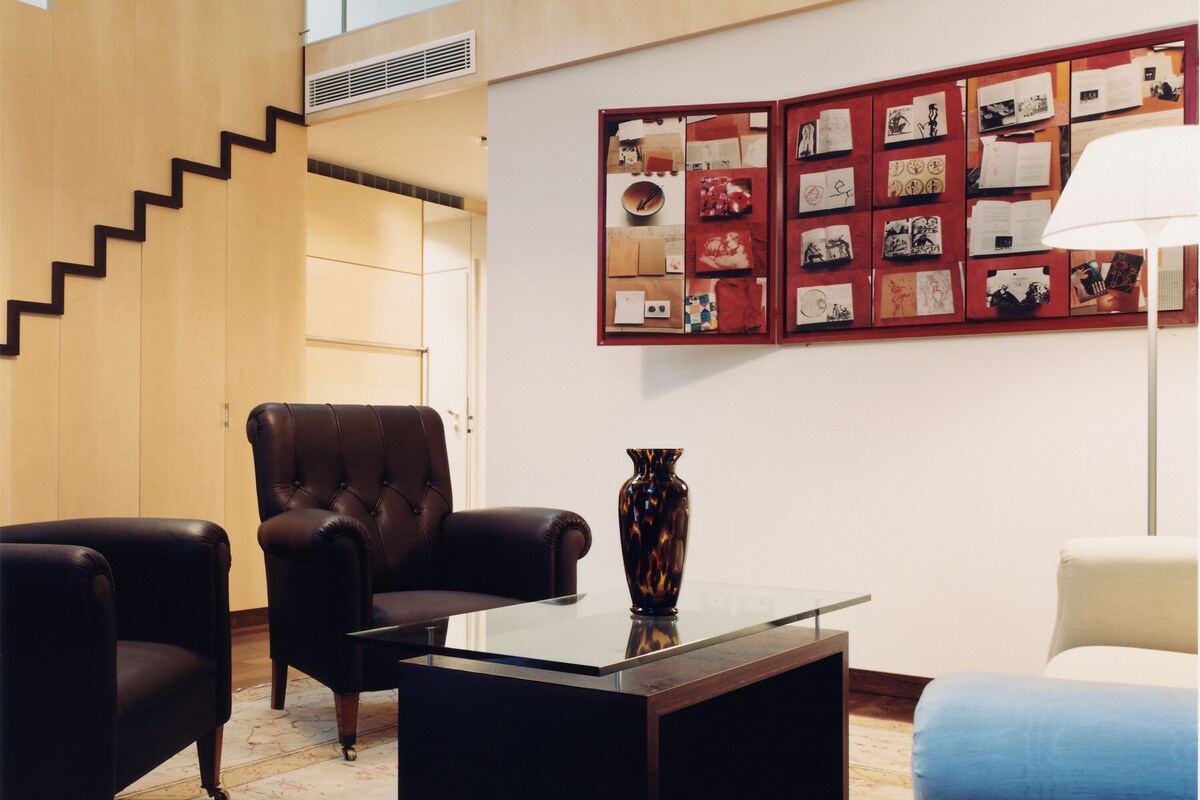 A modern living room with a staircase, leather armchairs, a glass coffee table, and a wall with framed pictures.