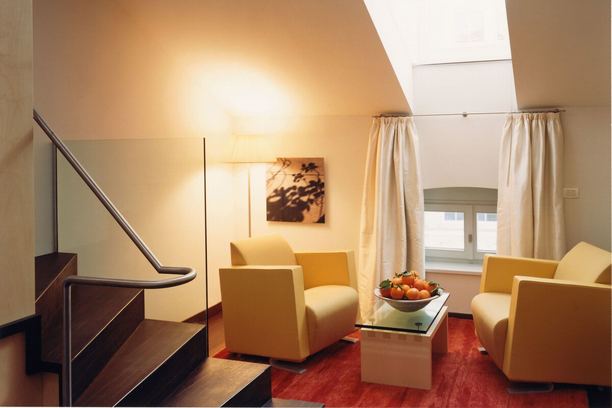 A modern living room with a staircase, two armchairs, a coffee table with a fruit bowl, a floor lamp, and a painting on the wall.