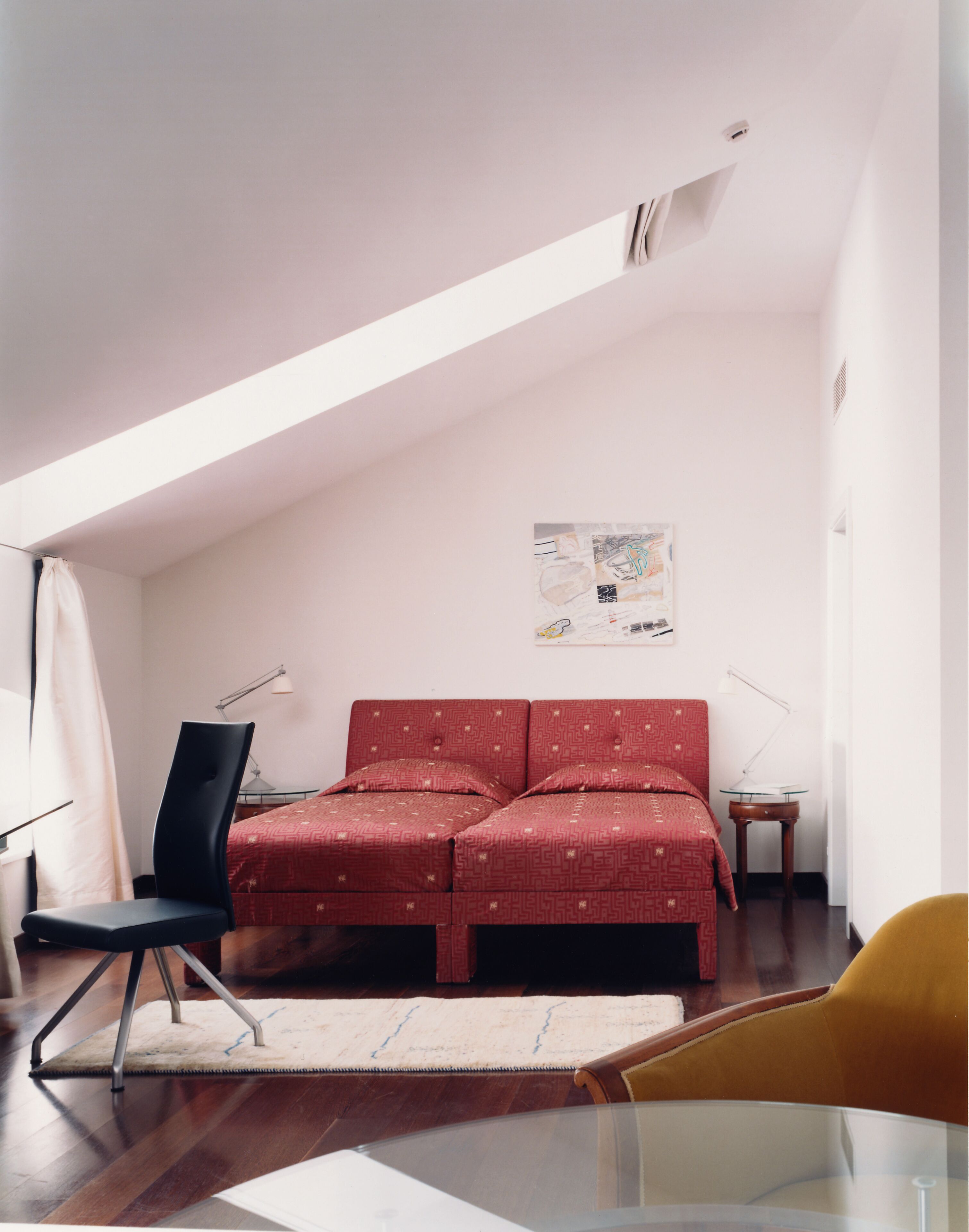 A bedroom with a sloped ceiling, a bed with patterned bedding, a desk with a chair, a small table, and a wall-mounted artwork.
