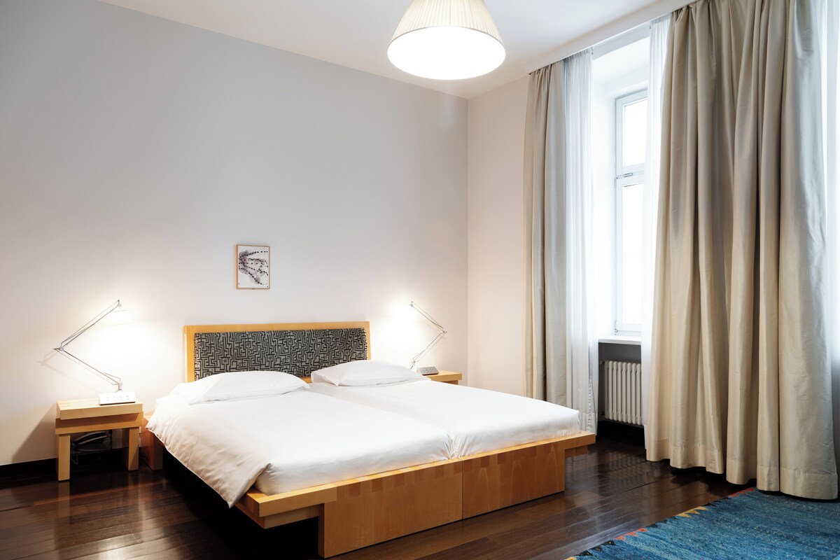 A modern bedroom with a wooden bed, two bedside tables, a floor lamp, a window with curtains, and a radiator.