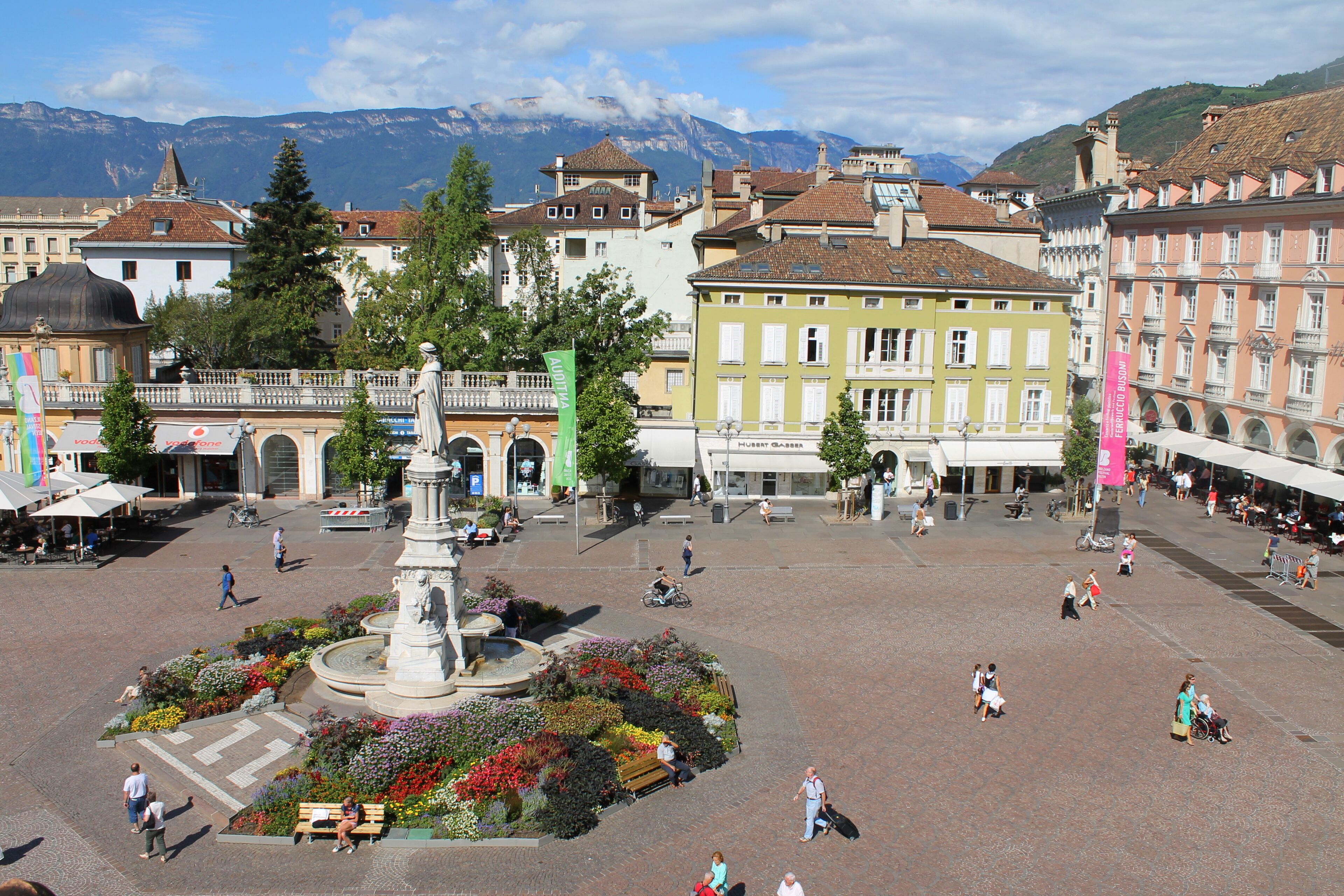 A public square with a central fountain, surrounded by historic buildings and a mountain backdrop.