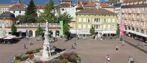 Una plaza pública con una fuente central, rodeada de edificios históricos y un telón de fondo montañoso.