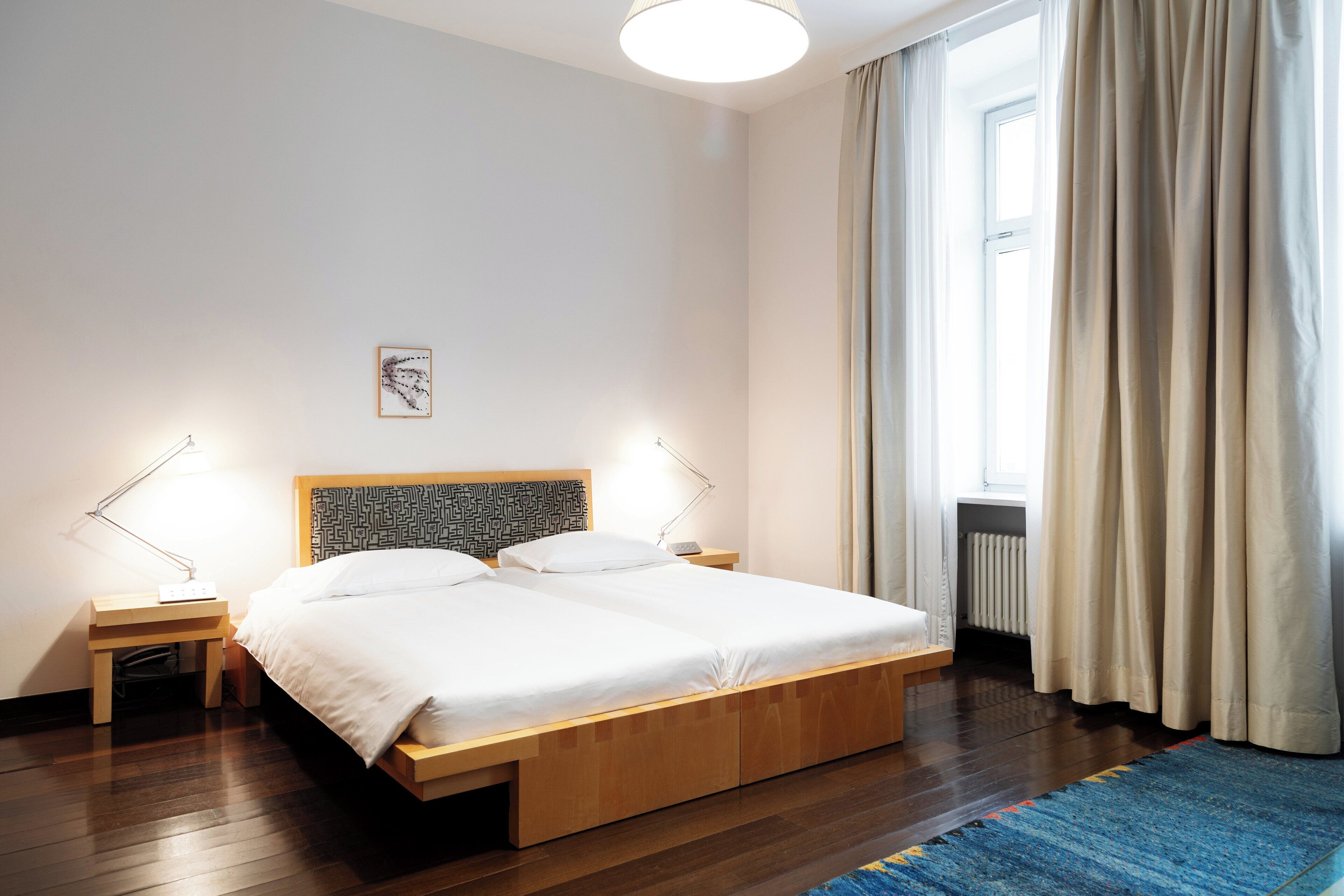 Un letto rifatto con cura, con struttura in legno e biancheria bianca, due comodini con lampade, un piccolo comodino con una lampada, una finestra con tende color beige e un tappeto blu sul pavimento.