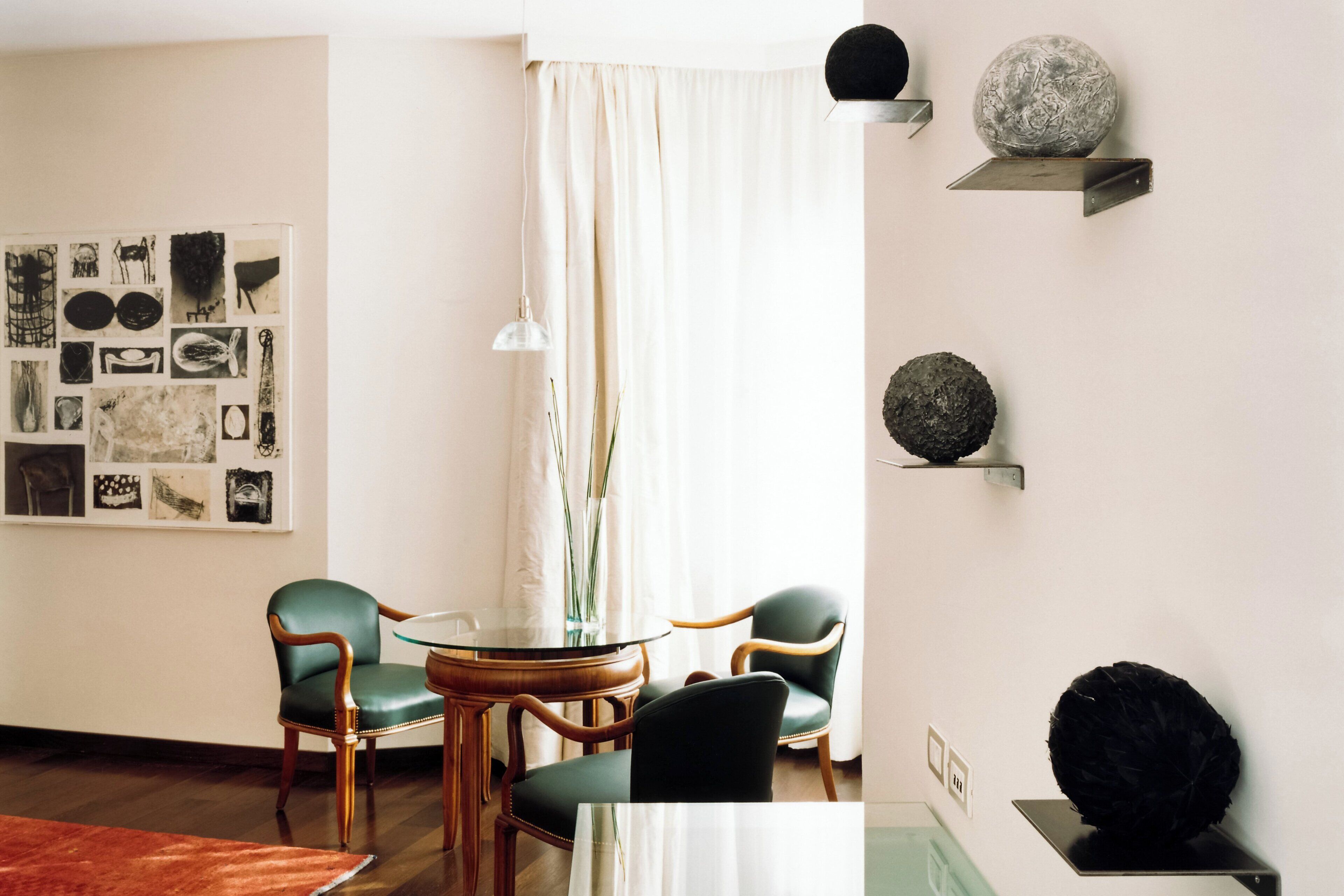 A modern living room with a round glass table, two green chairs, a wooden coffee table, a vase with long stems, and a wall-mounted shelf with decorative spheres.