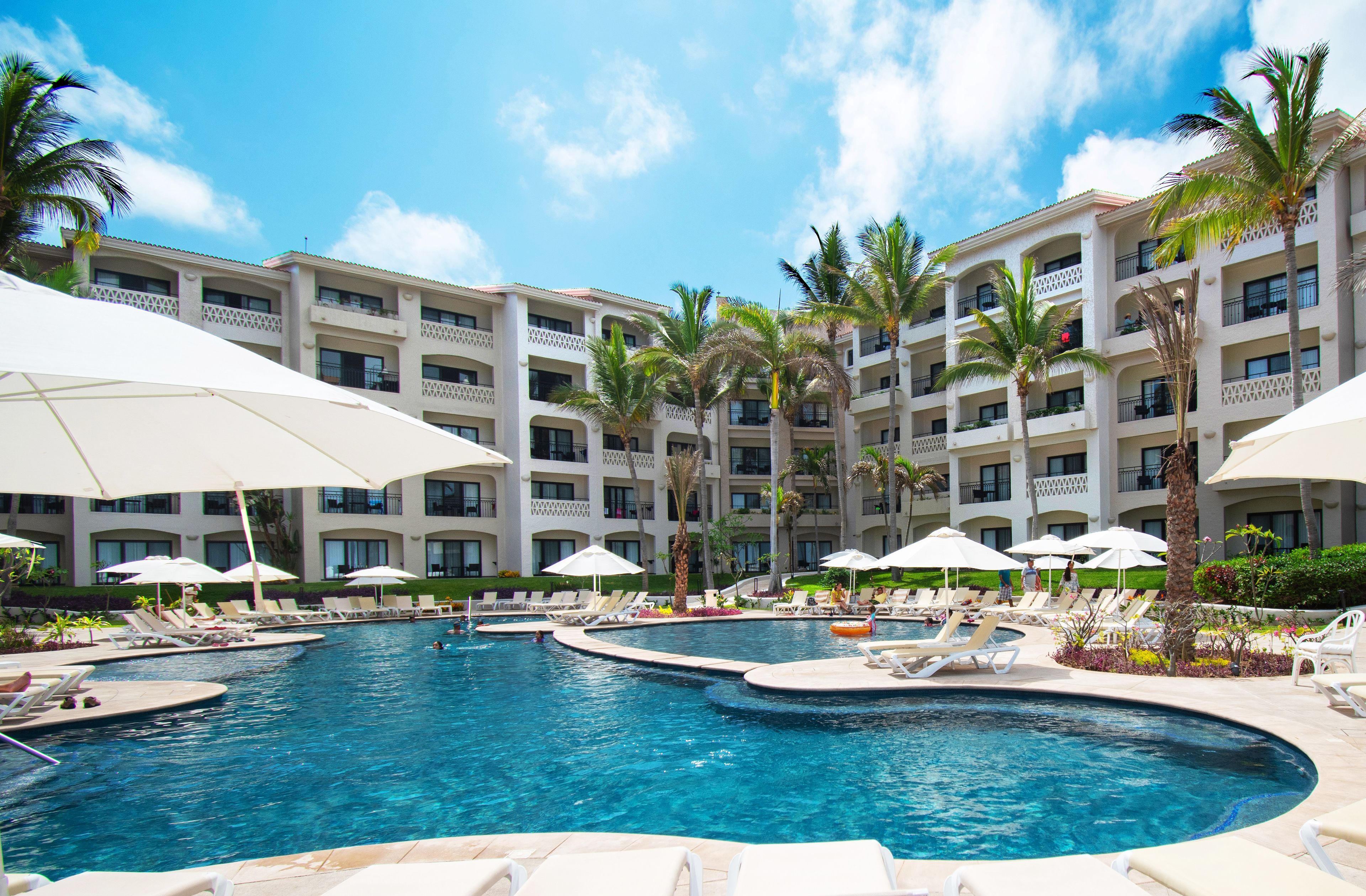A hotel with multiple swimming pools, palm trees, and white umbrellas.