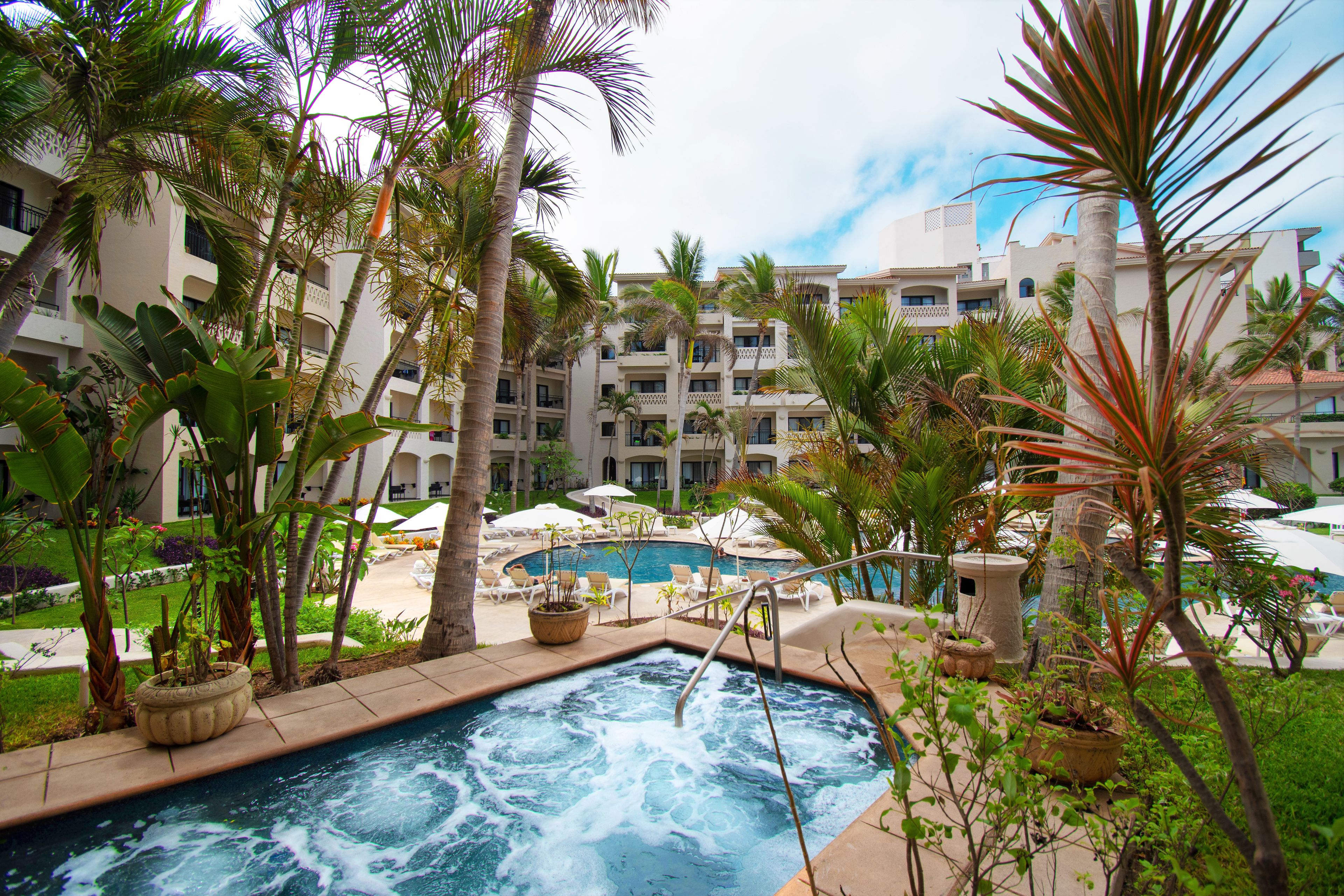 Zona de piscina de hotel con jacuzzi, rodeada de palmeras y sillones de descanso.