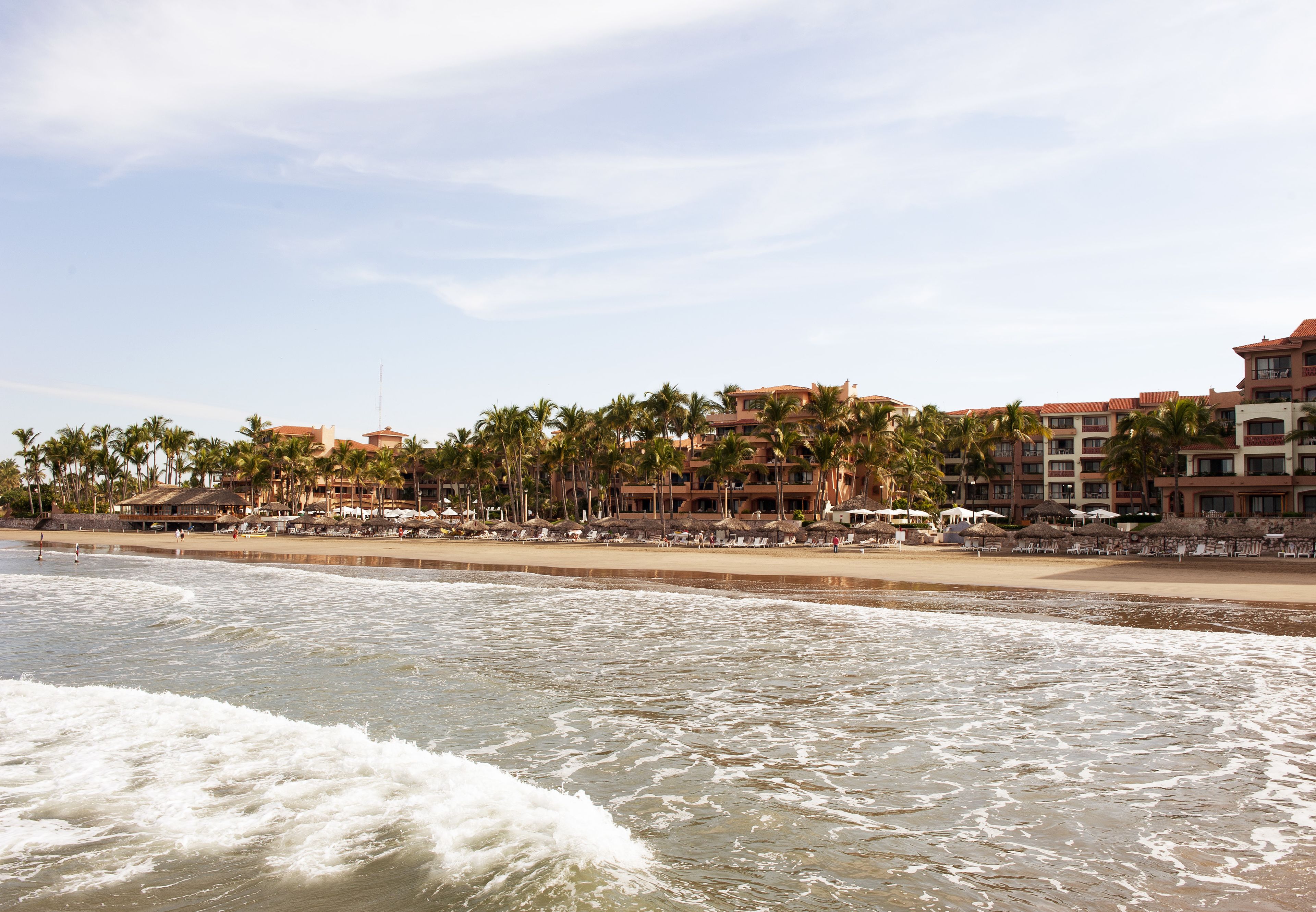 Um hotel à beira-mar com vários andares, palmeiras e guarda-sóis de praia.