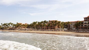 Um hotel à beira-mar com vários andares, palmeiras e guarda-sóis de praia.