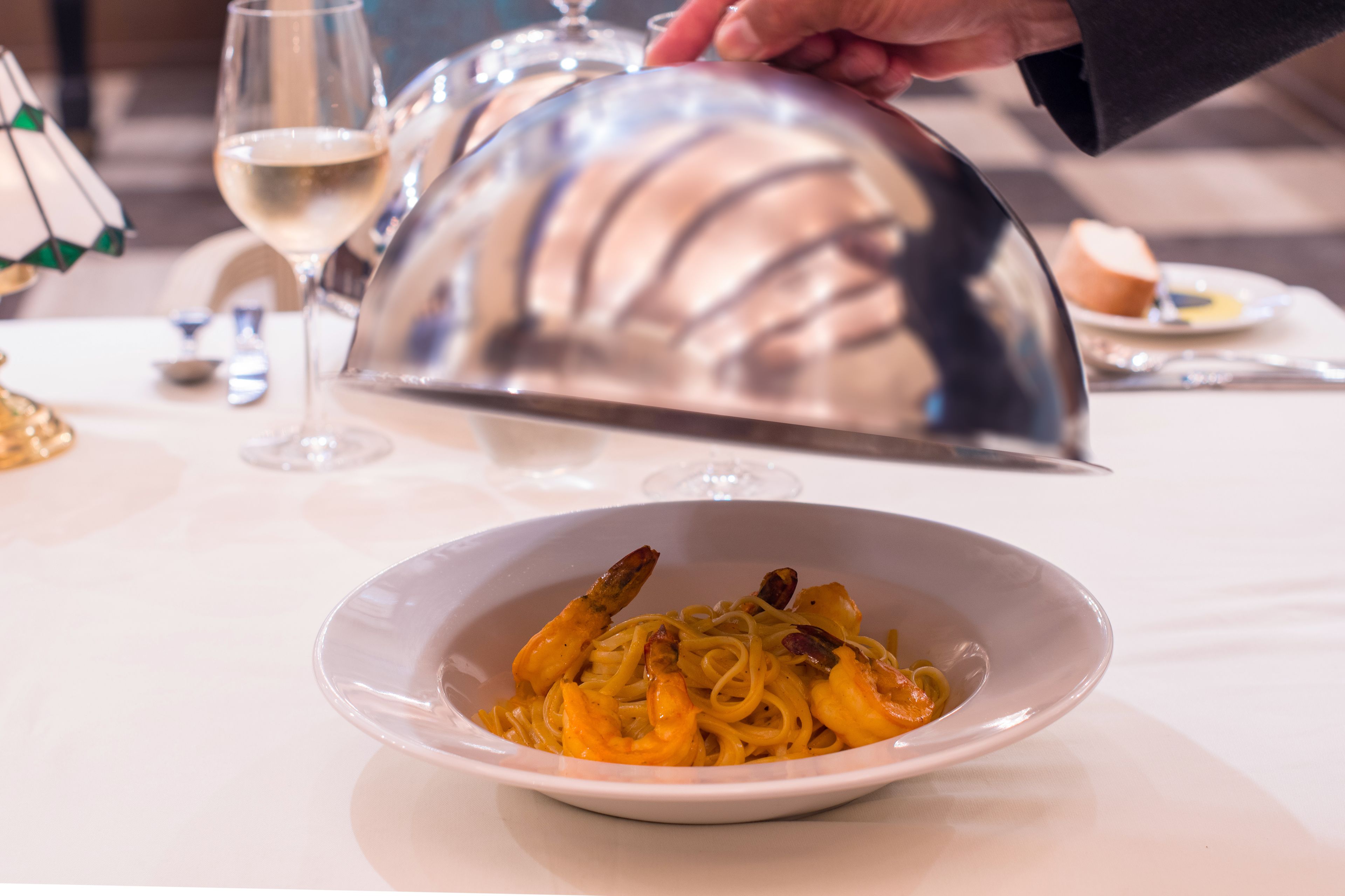 A white table with a plate of pasta and shrimp, a wine glass, and a decorative lamp.