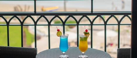 A balcony with two wicker chairs and a round table, overlooking a beach with palm trees and the ocean.
