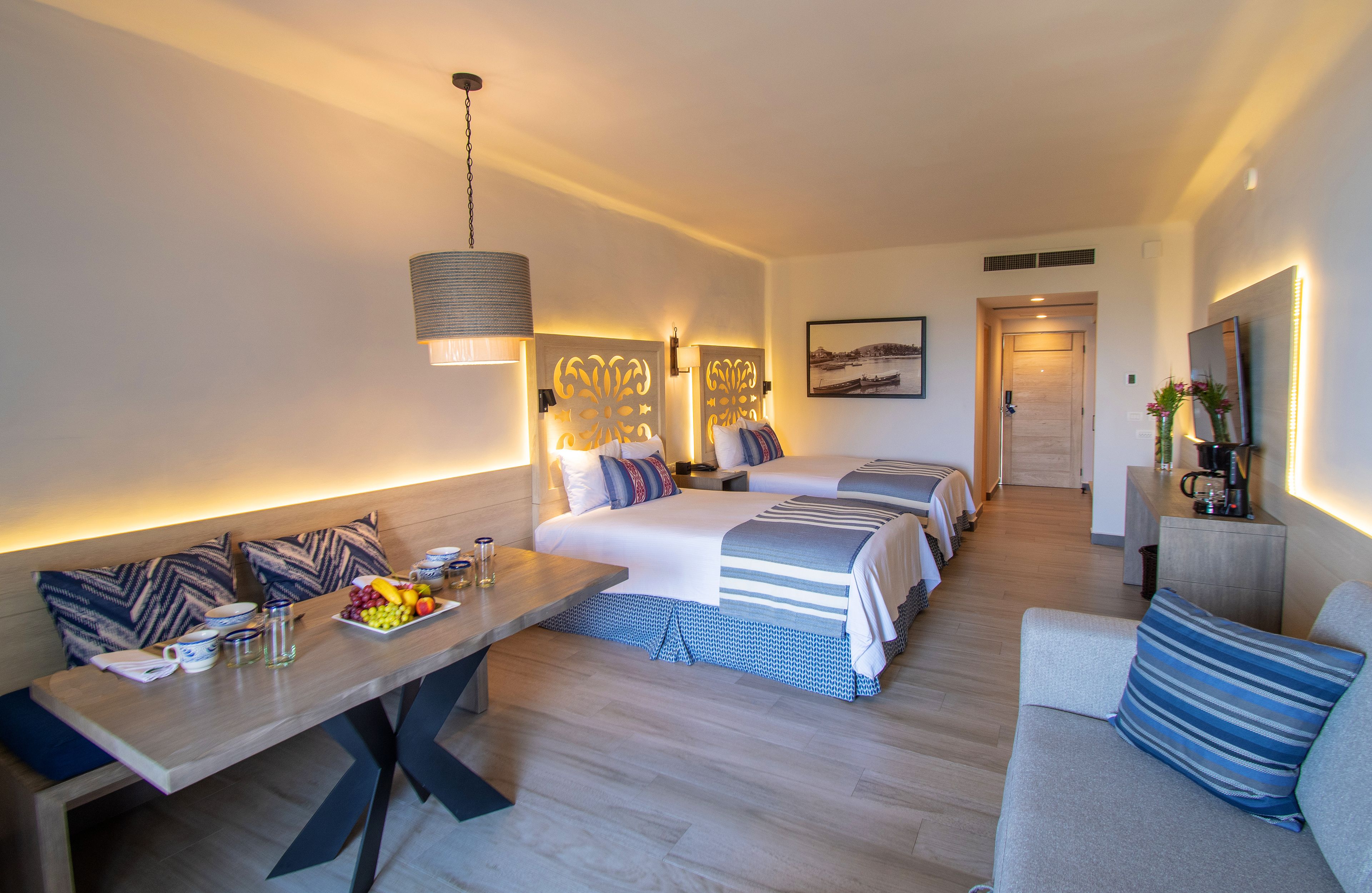 A hotel room with a dining table, two beds, a sofa, and a small kitchen area.