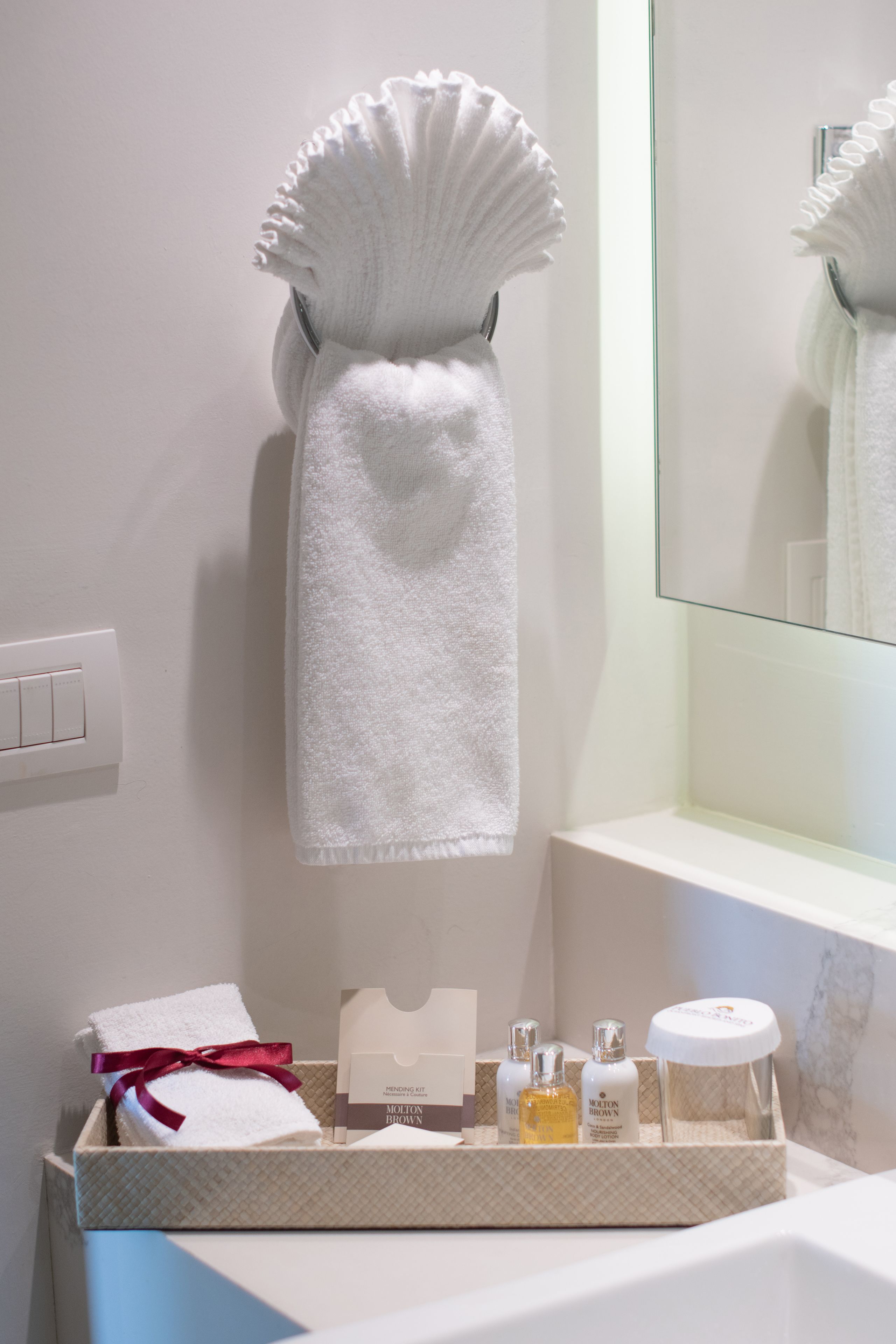 Une salle de bain avec une serviette blanche suspendue à un porte-serviettes, un miroir, un plateau contenant des articles de toilette et un interrupteur sur le mur.