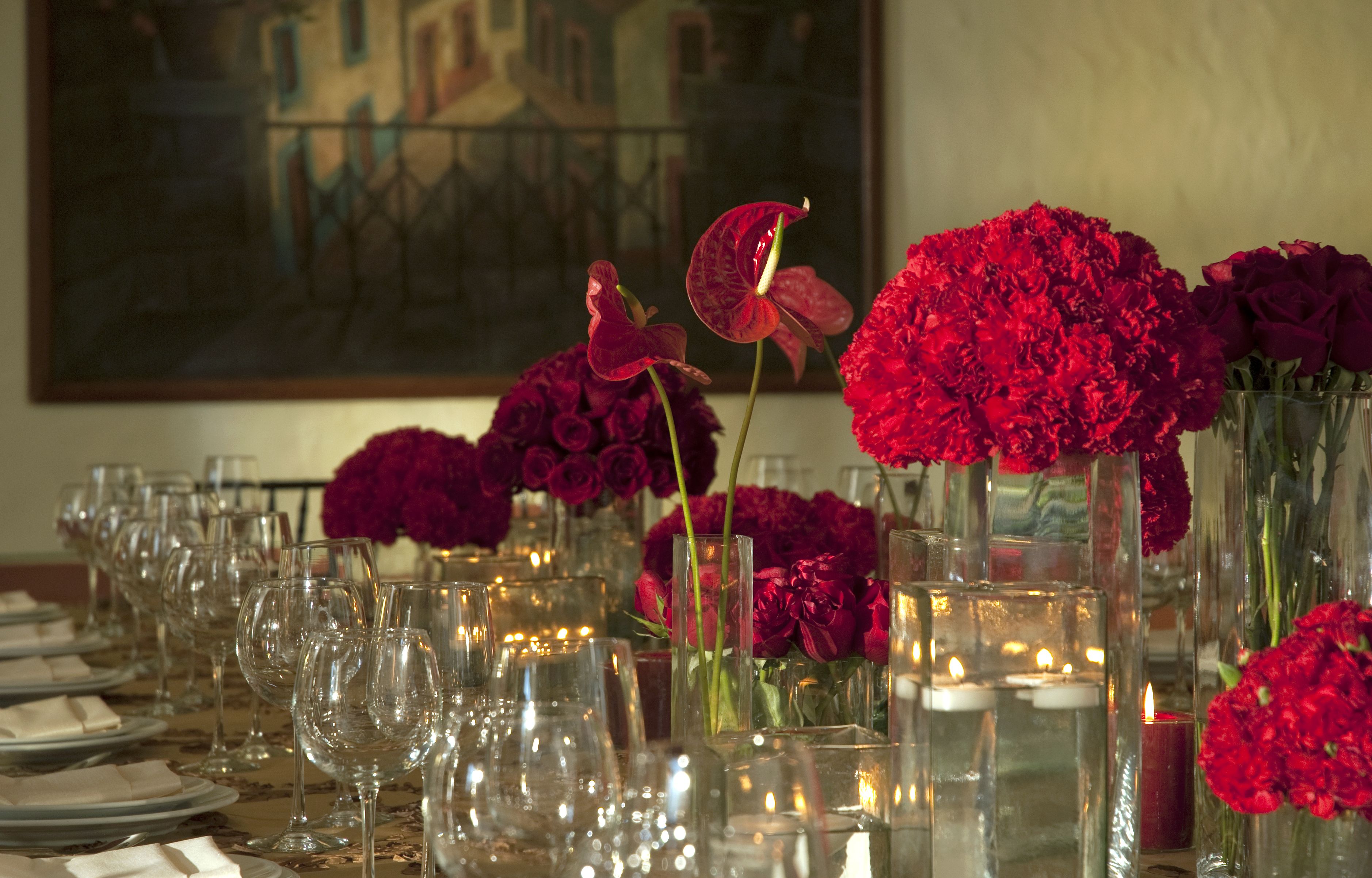 A table set for dining with multiple glasses and plates, accompanied by vases of red flowers and candles.