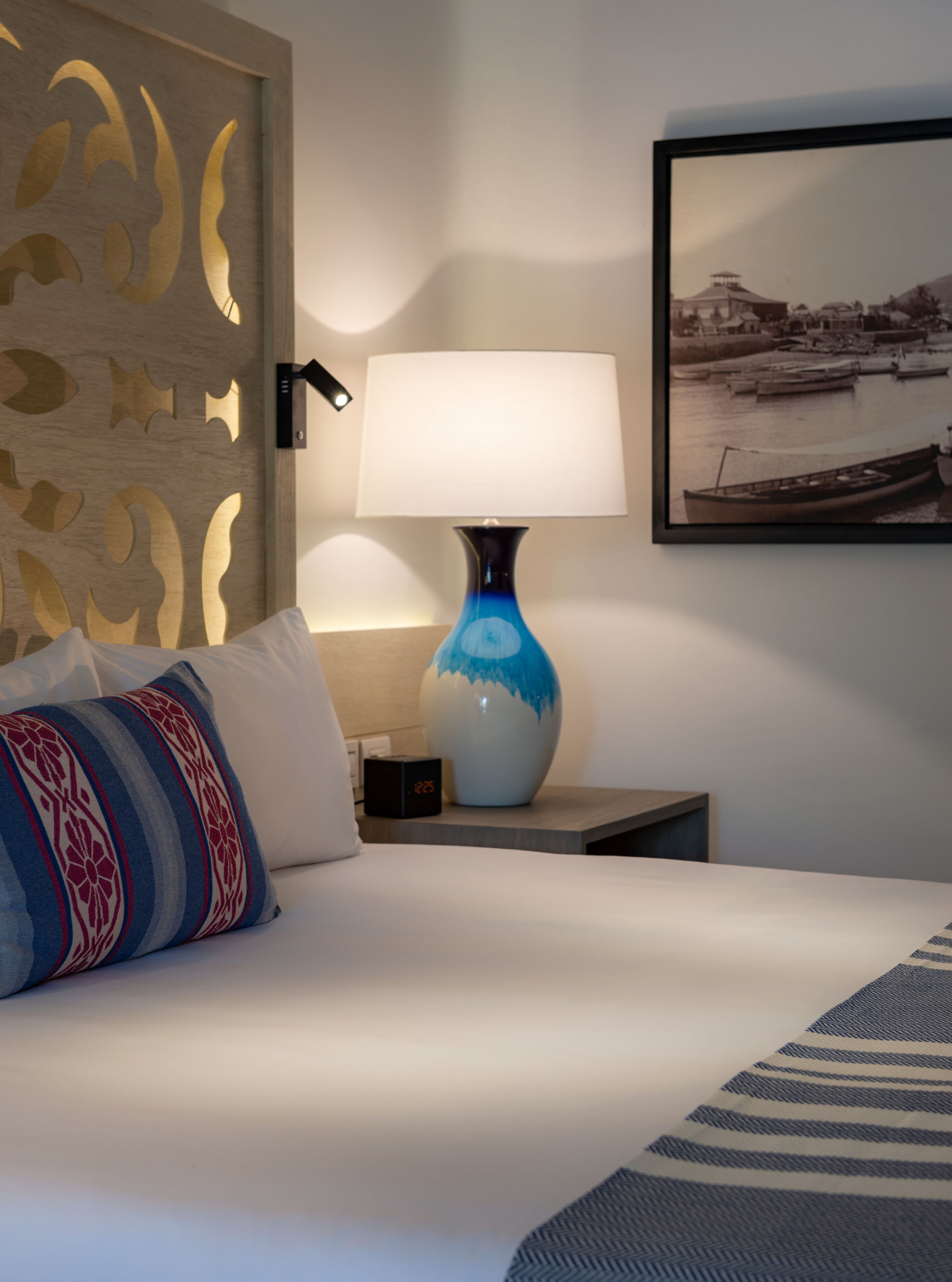 A neatly made bed with a white comforter and a blue and white patterned pillow. A blue lamp with a white shade is on a bedside table, and a framed photograph hangs on the wall.