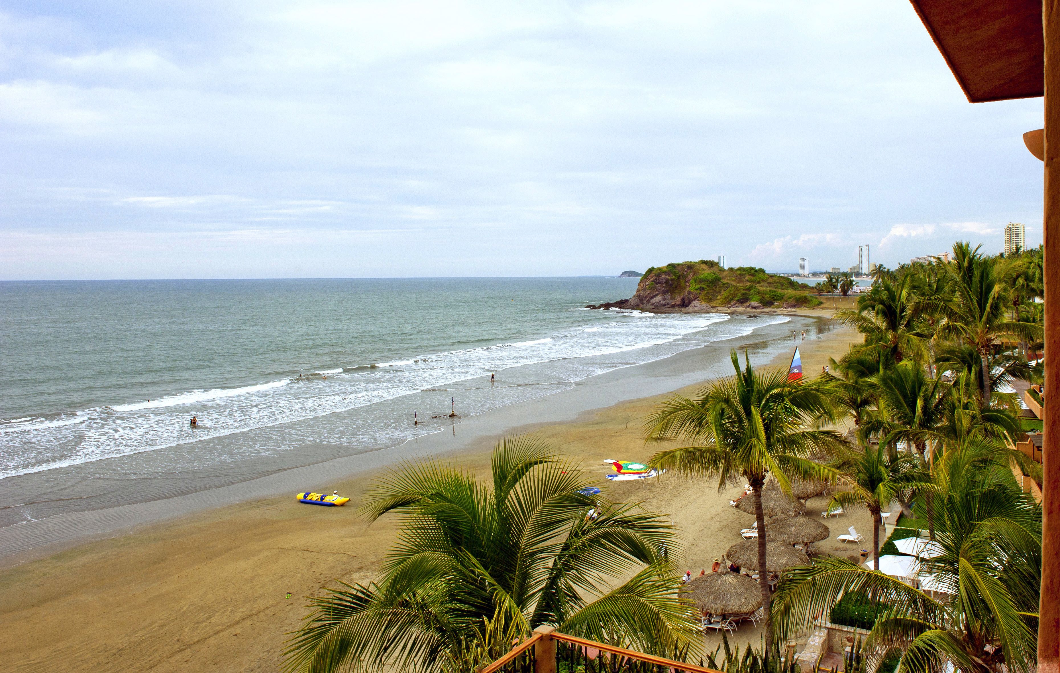 A beach with a view of the ocean, palm trees, and beach umbrellas.