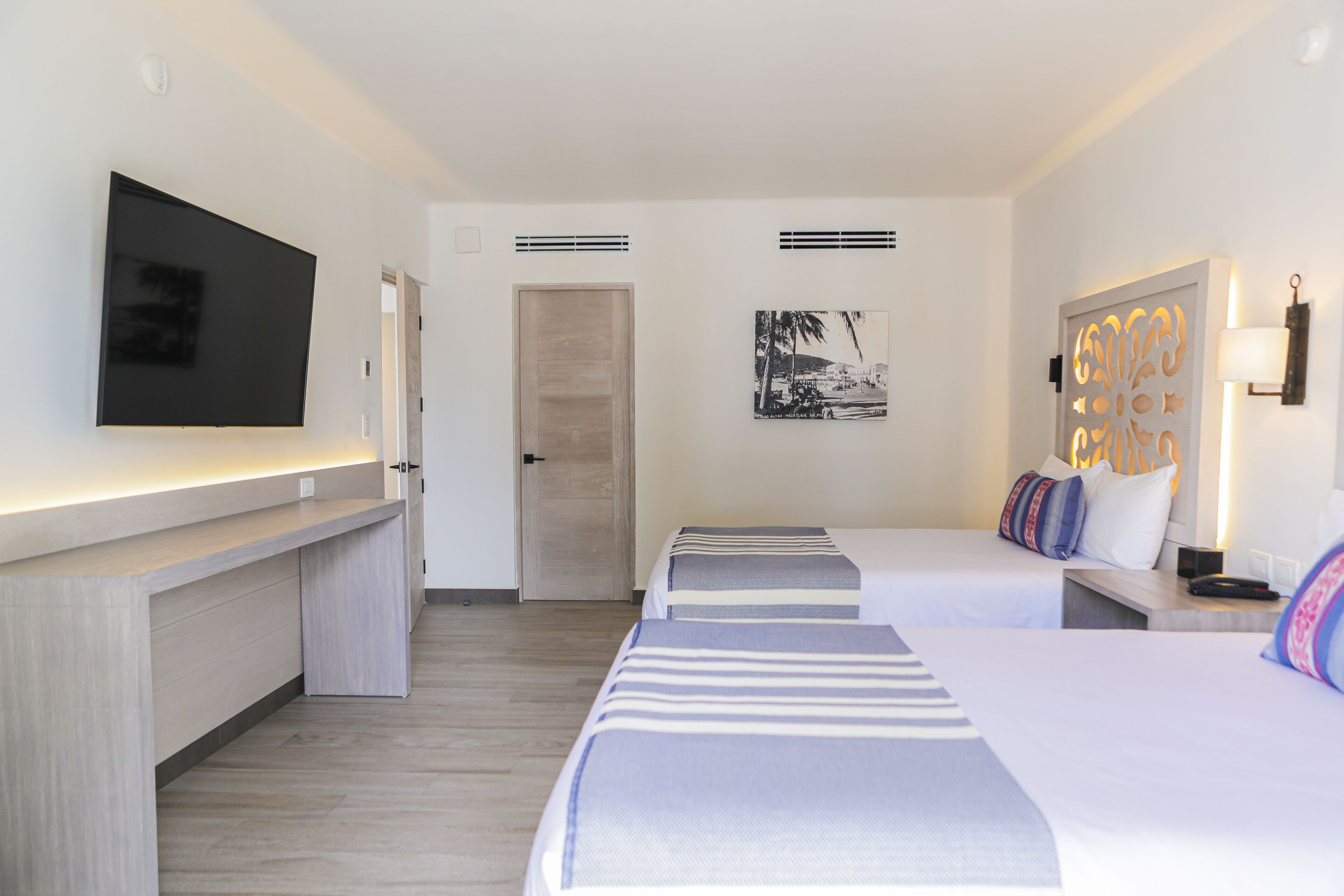 A hotel room with a flat-screen TV mounted on the wall, a desk with a lighted surface, two beds with striped bedding, a door, a wall-mounted artwork, and a decorative wall light.