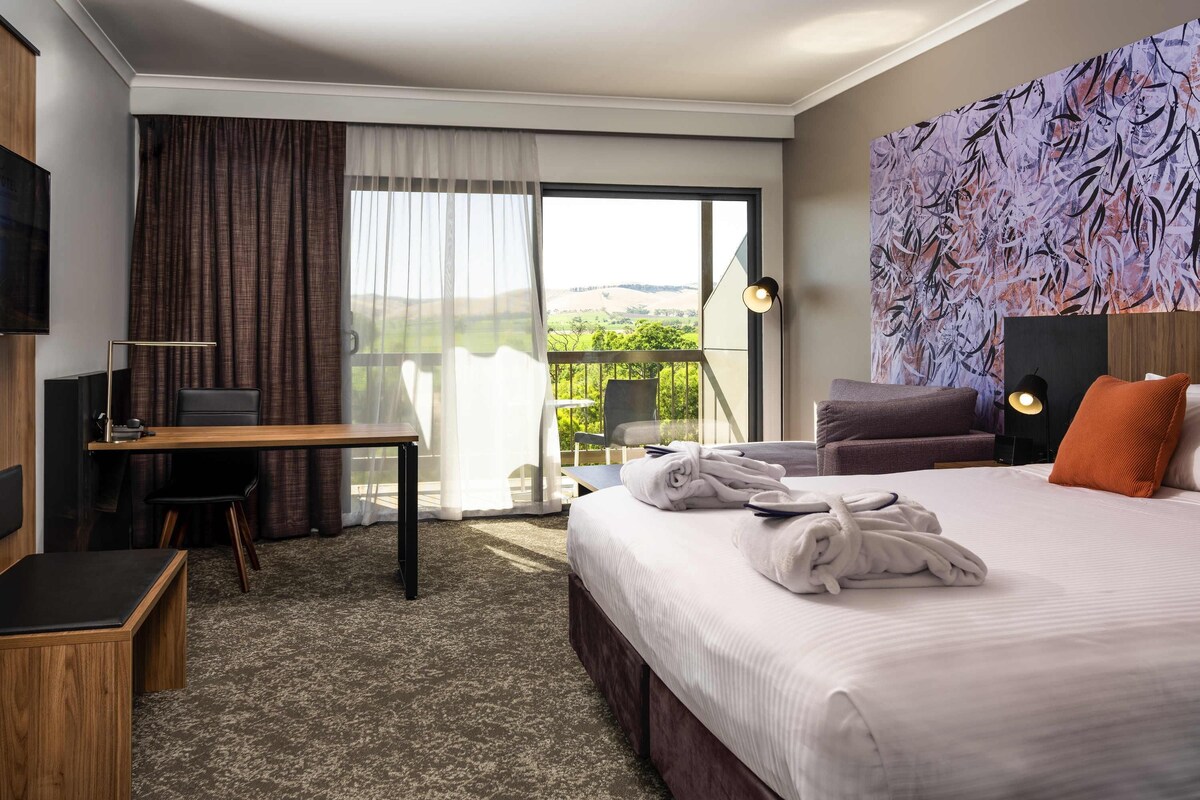 Habitación de hotel con cama, escritorio, silla, televisor y balcón con vistas.