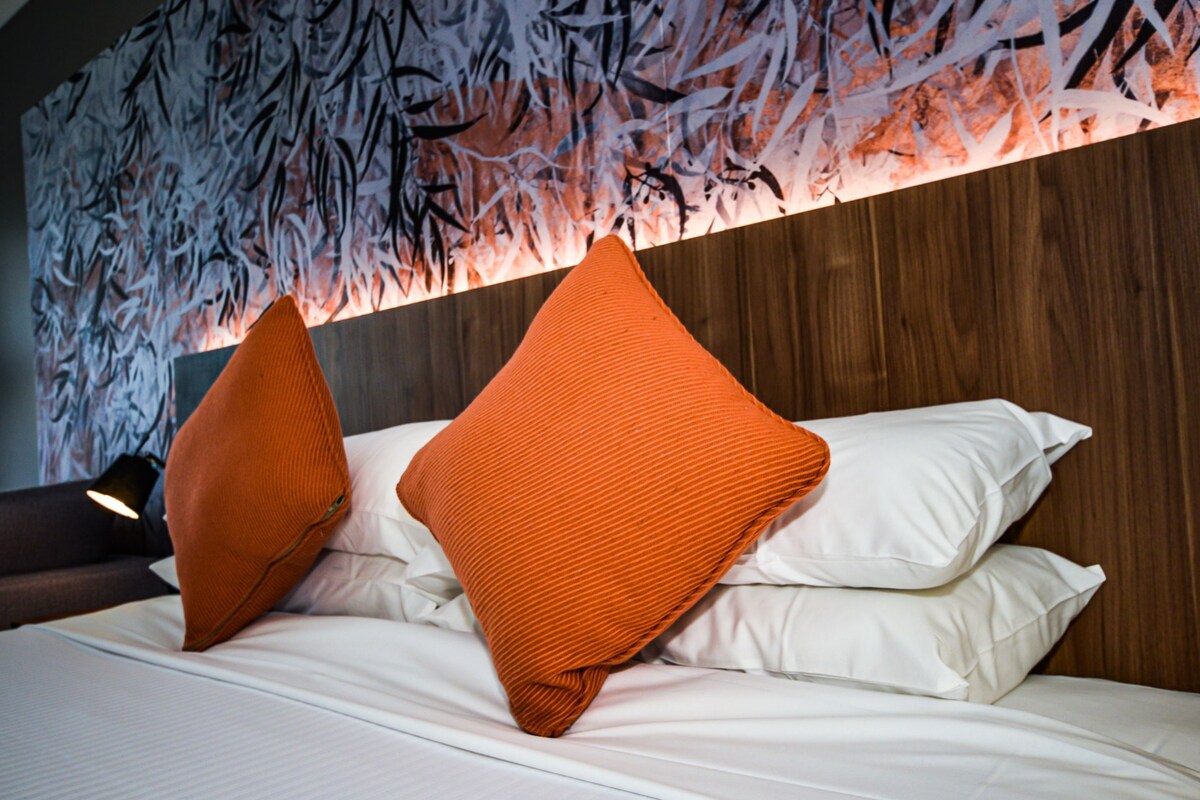 Habitación de hotel con una cama, dos almohadas naranjas, ropa de cama blanca y una pared con diseño de hojas.