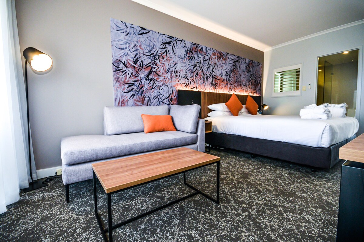 Una habitación de hotel con un sofá gris, una mesa de centro de madera, una cama con ropa blanca y almohadas naranjas, un cuadro abstracto grande en la pared y suelo alfombrado.