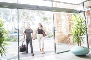 Un hombre y una mujer caminan tomados de la mano a través de una puerta de cristal, con una planta en maceta y una jardinera azul grande al frente.