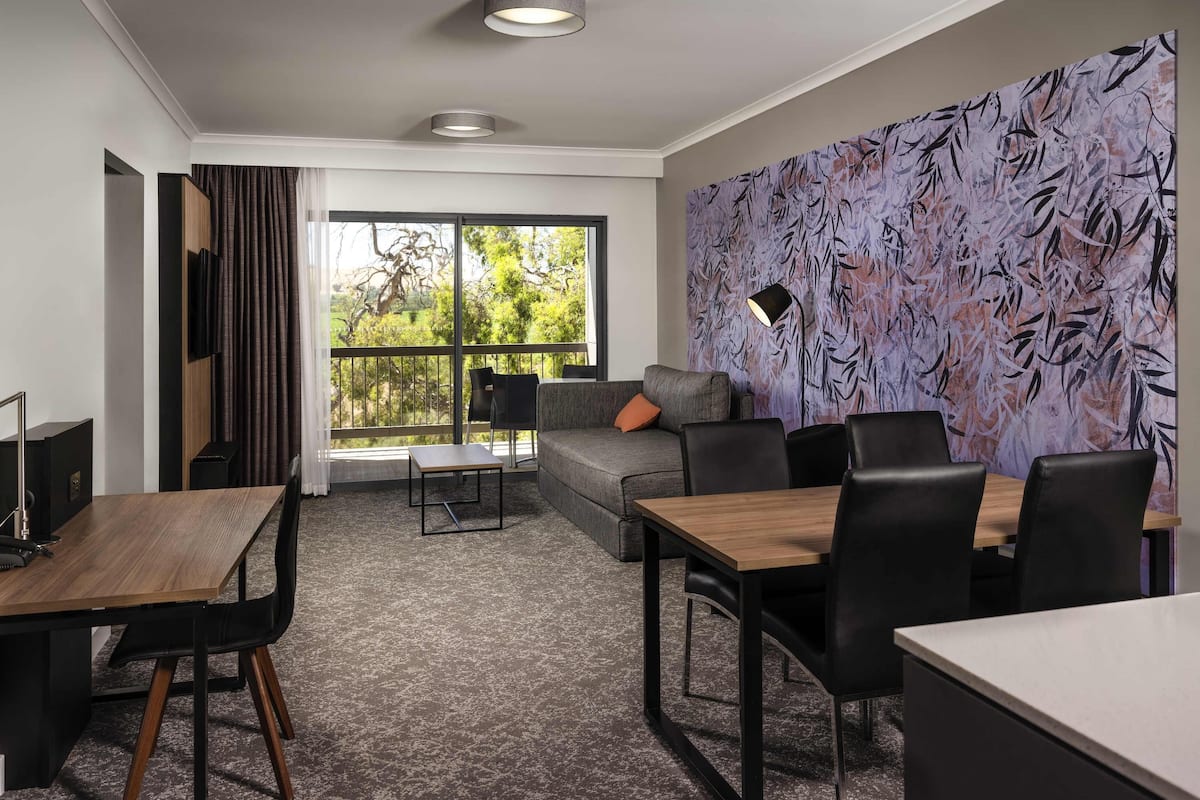 A modern hotel room with a dining area, a sofa, and a large window with a view of greenery.