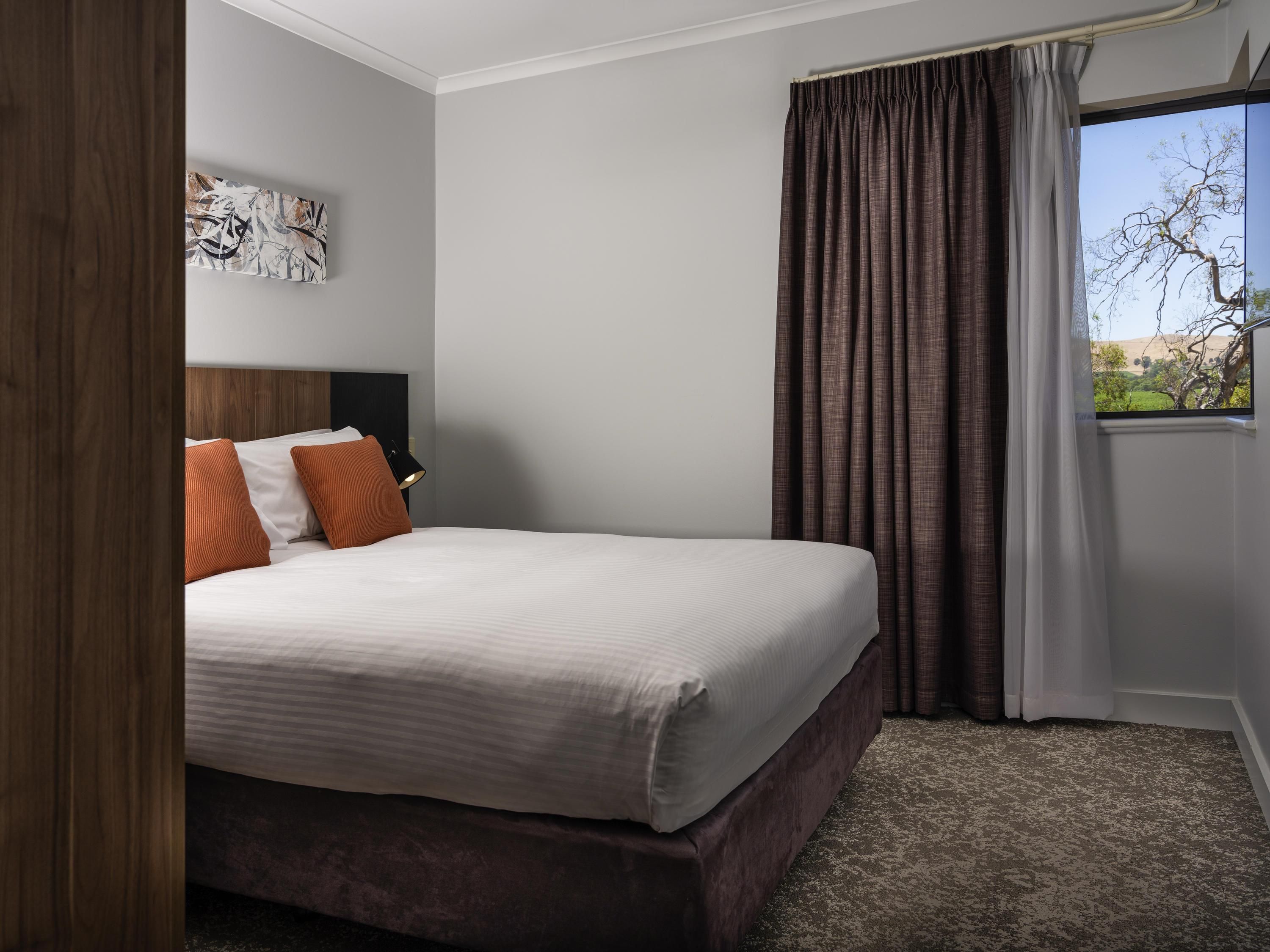 A hotel room with a large bed, a headboard, a nightstand, a lamp, a window with curtains, and a view of trees and hills.
