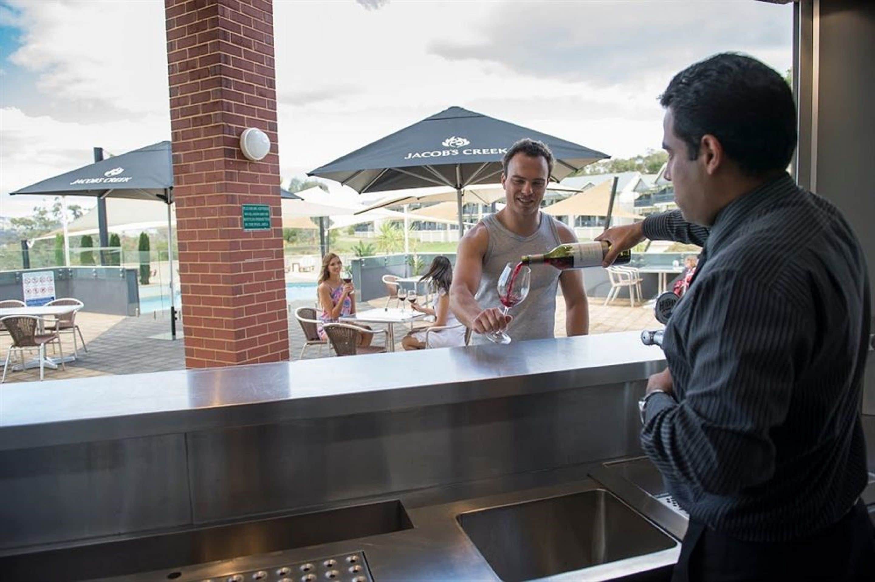 A man pouring a drink at a bar with outdoor seating and umbrellas labeled "Jacob's Creek".