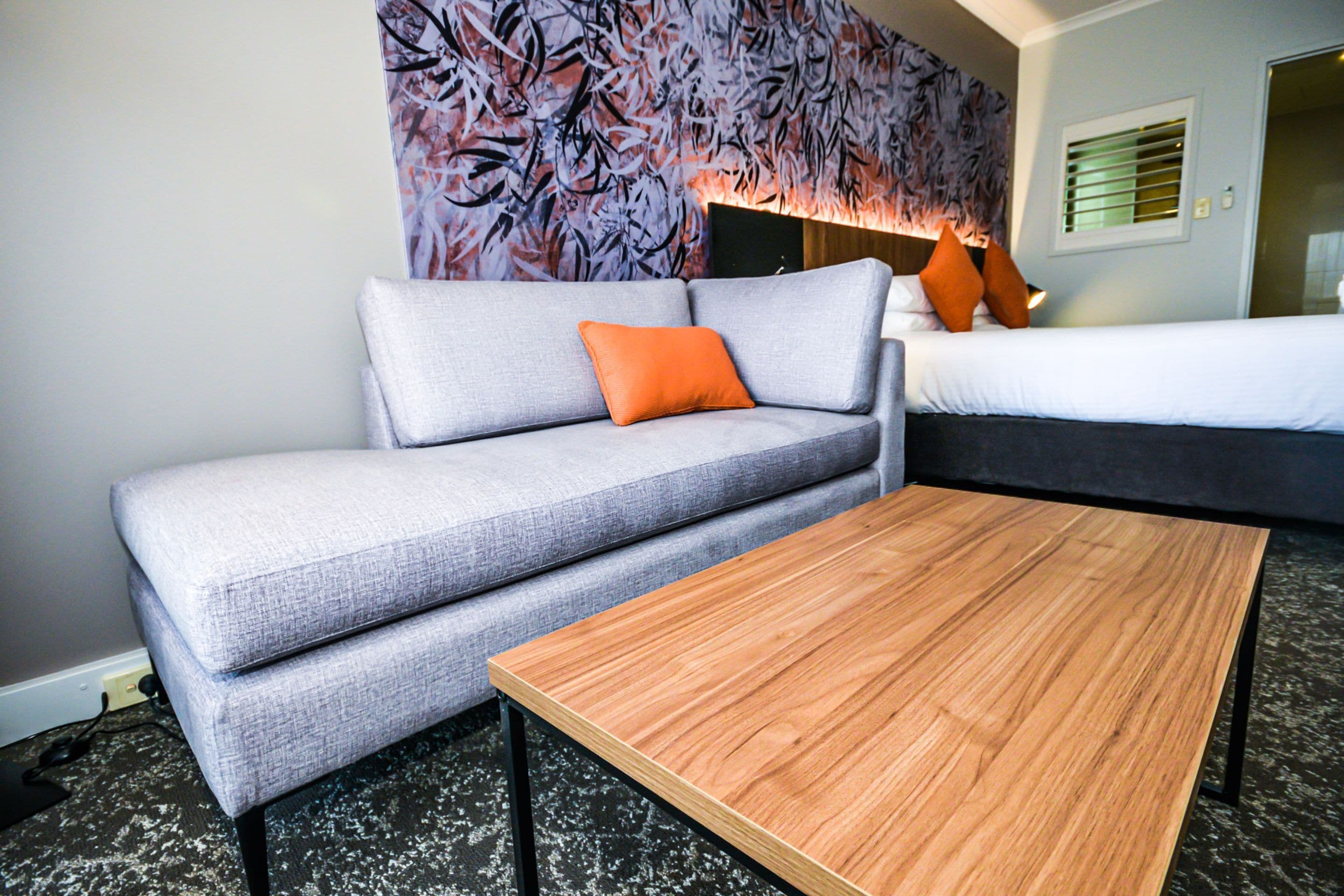 A modern hotel room with a grey sofa, an orange pillow, a wooden coffee table, and a bed with a dark headboard.