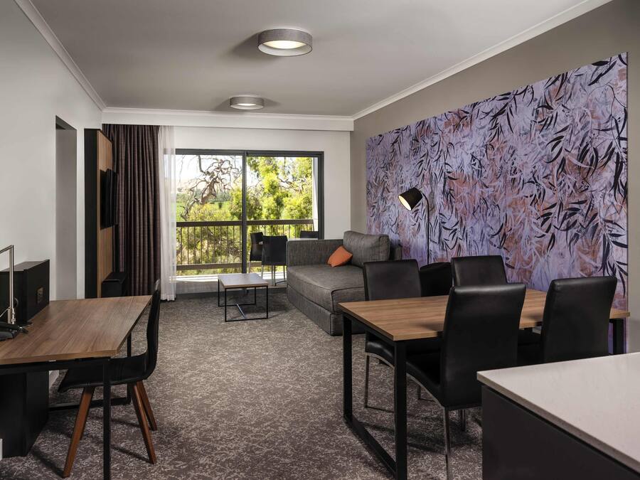 A modern hotel room with a dining area, a sofa, and a large window with a view of greenery.