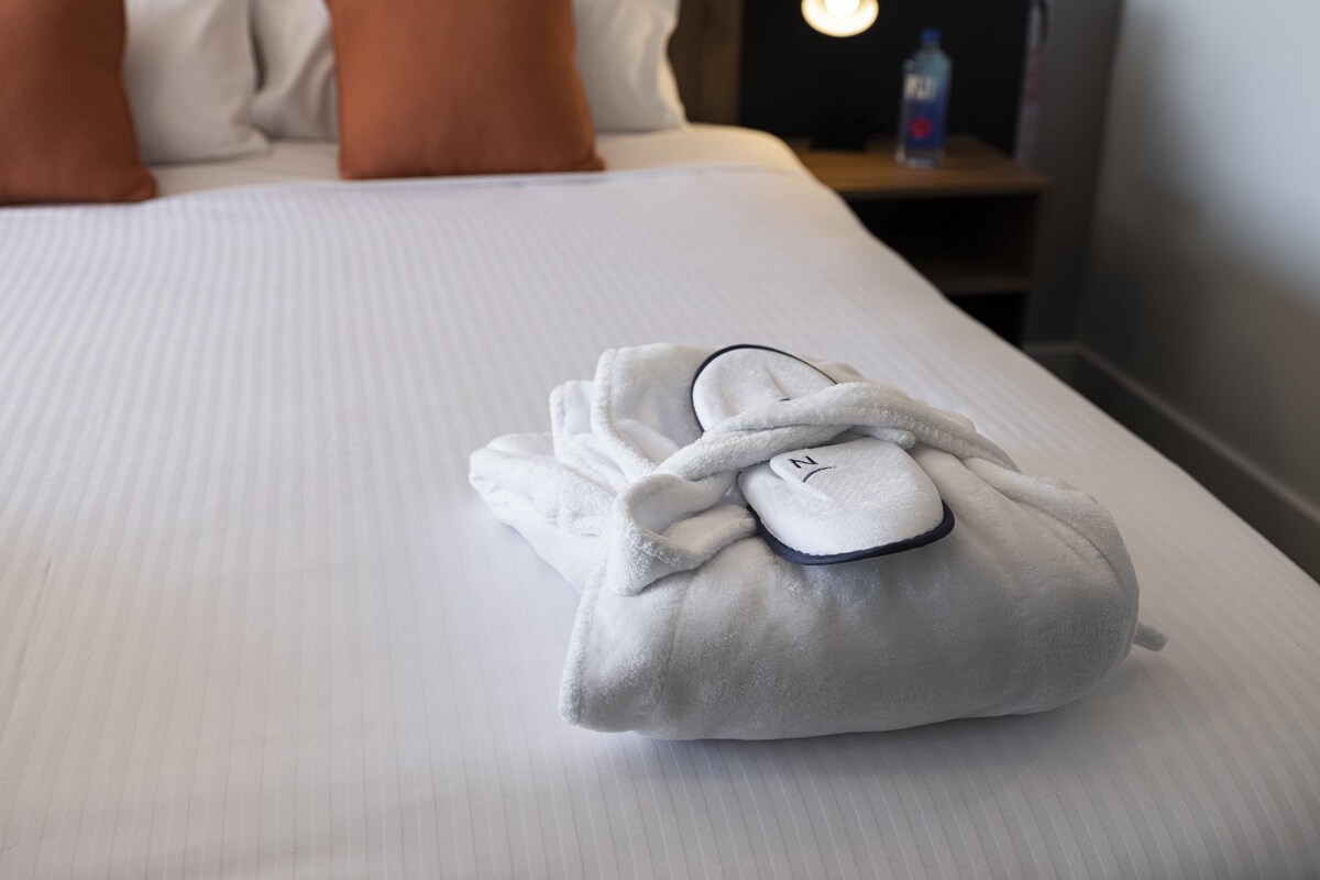 Una cama bien hecha con ropa blanca y dos almohadas naranjas. Una toalla blanca y zapatillas están sobre la cama.