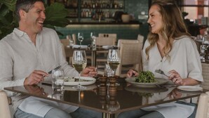 Una pareja cenando en una mesa con copas de vino y platos de comida, en un restaurante con barra y estantes llenos de botellas al fondo.