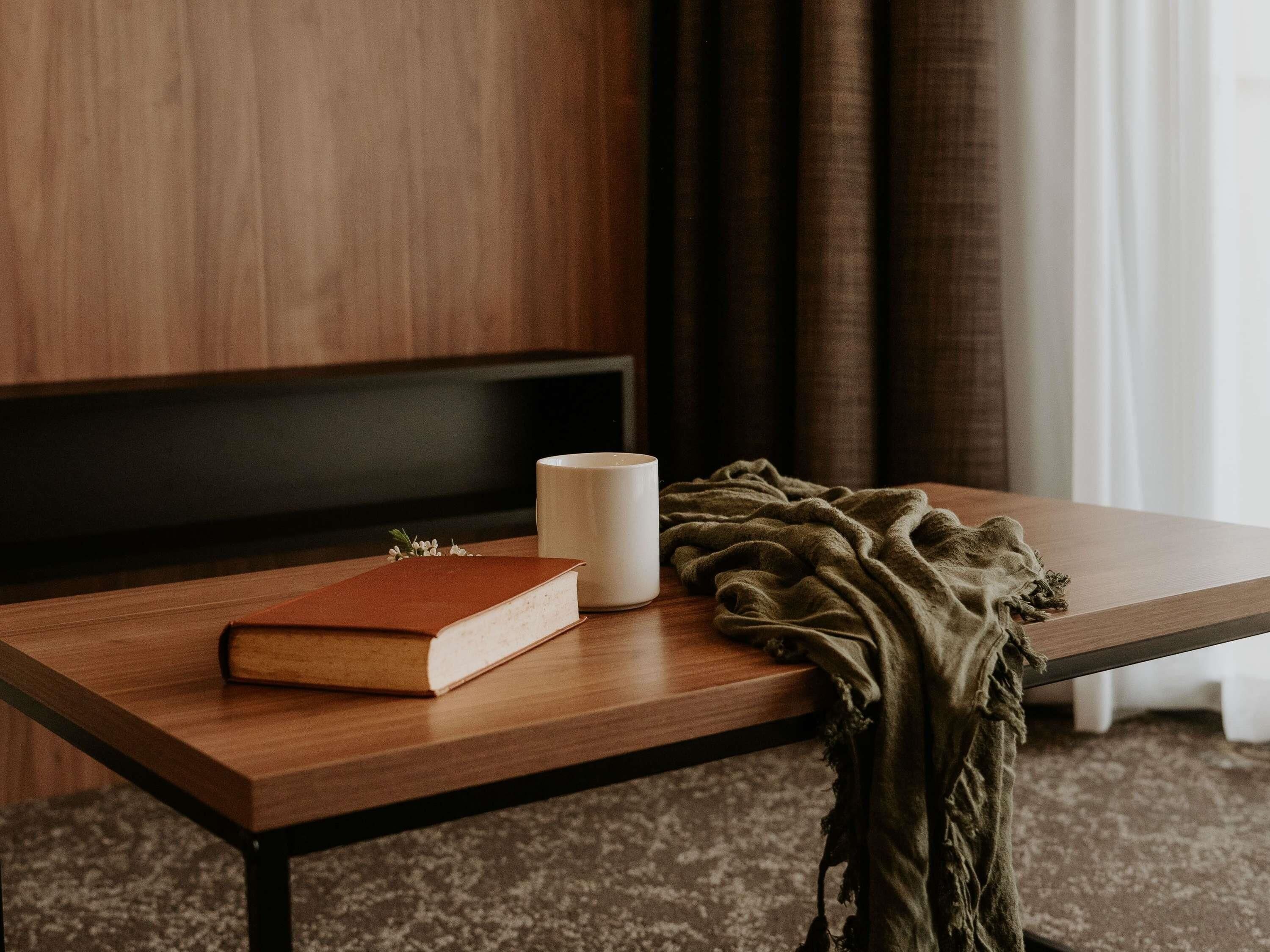 A wooden table with a book, a white mug, and a green blanket.