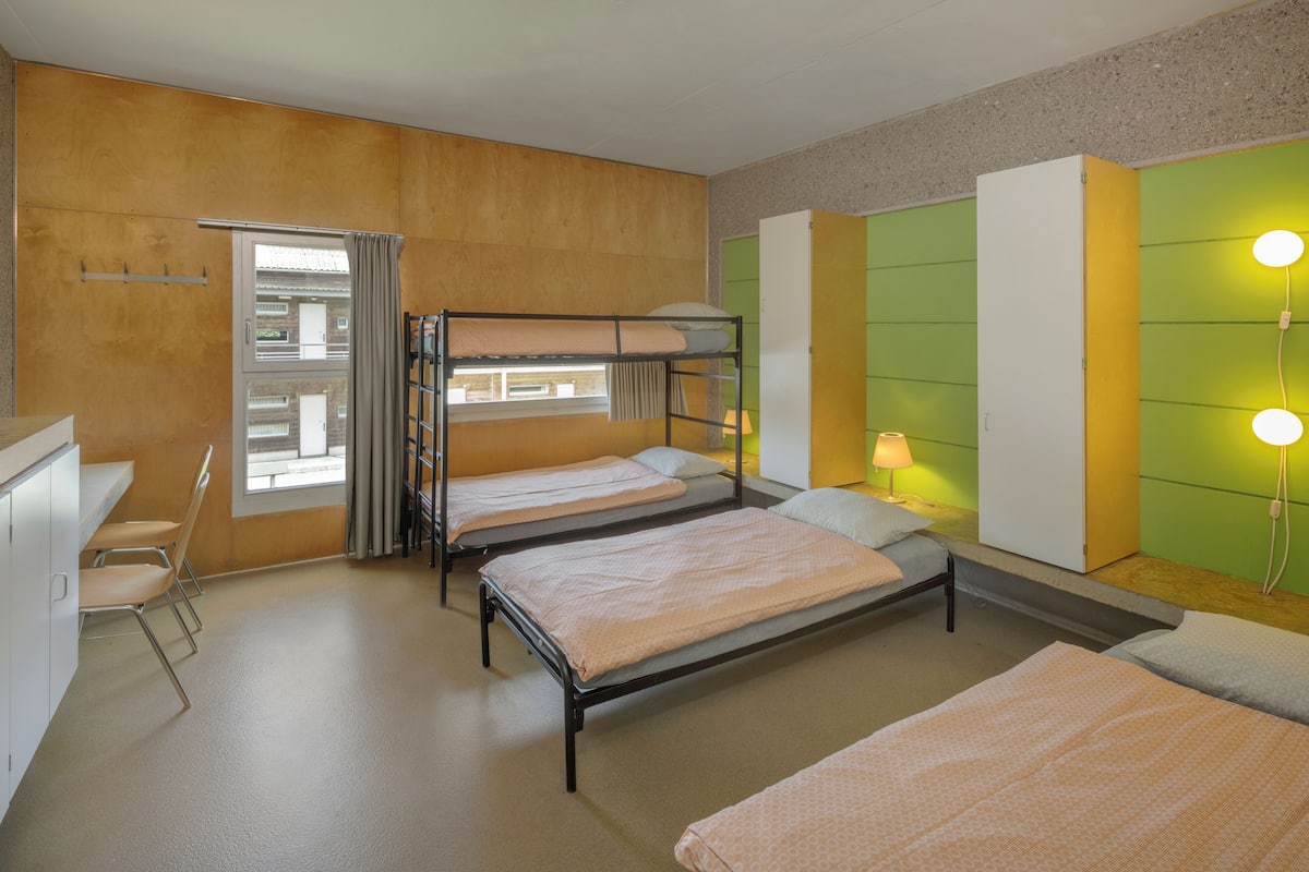 A dormitory room with bunk beds, a desk, chairs, and a window with curtains.