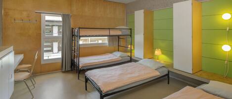 A dormitory room with bunk beds, a desk, chairs, and a window with curtains.