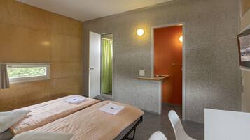 A modern hotel room with two beds, a small desk, and a shower area.