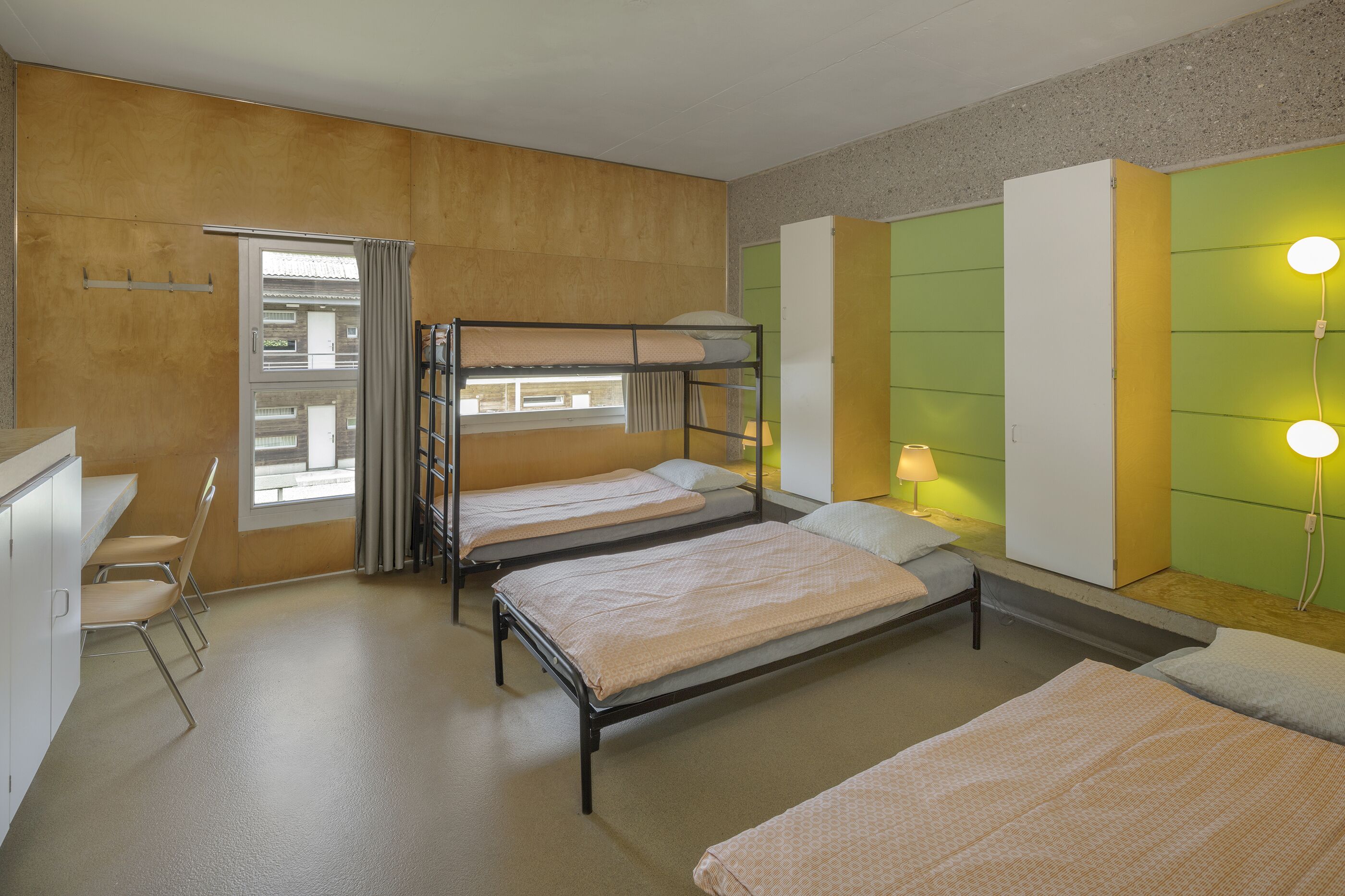 A dormitory room with bunk beds, a desk, chairs, and a window with curtains.