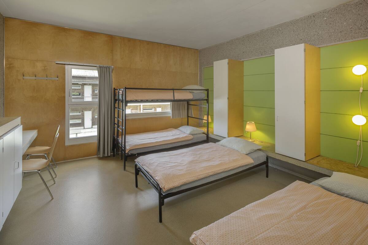 A dormitory room with bunk beds, a desk, chairs, and a window with curtains.