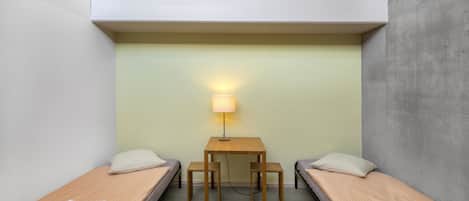Una habitación pequeña y funcional con dos camas individuales, una mesita y una lámpara.