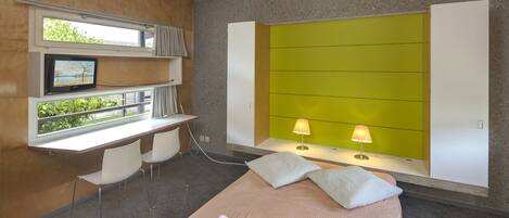 Habitación de hotel moderna con cama, dos sillas, un escritorio con computadora, televisor y ventana con vista a zonas verdes.