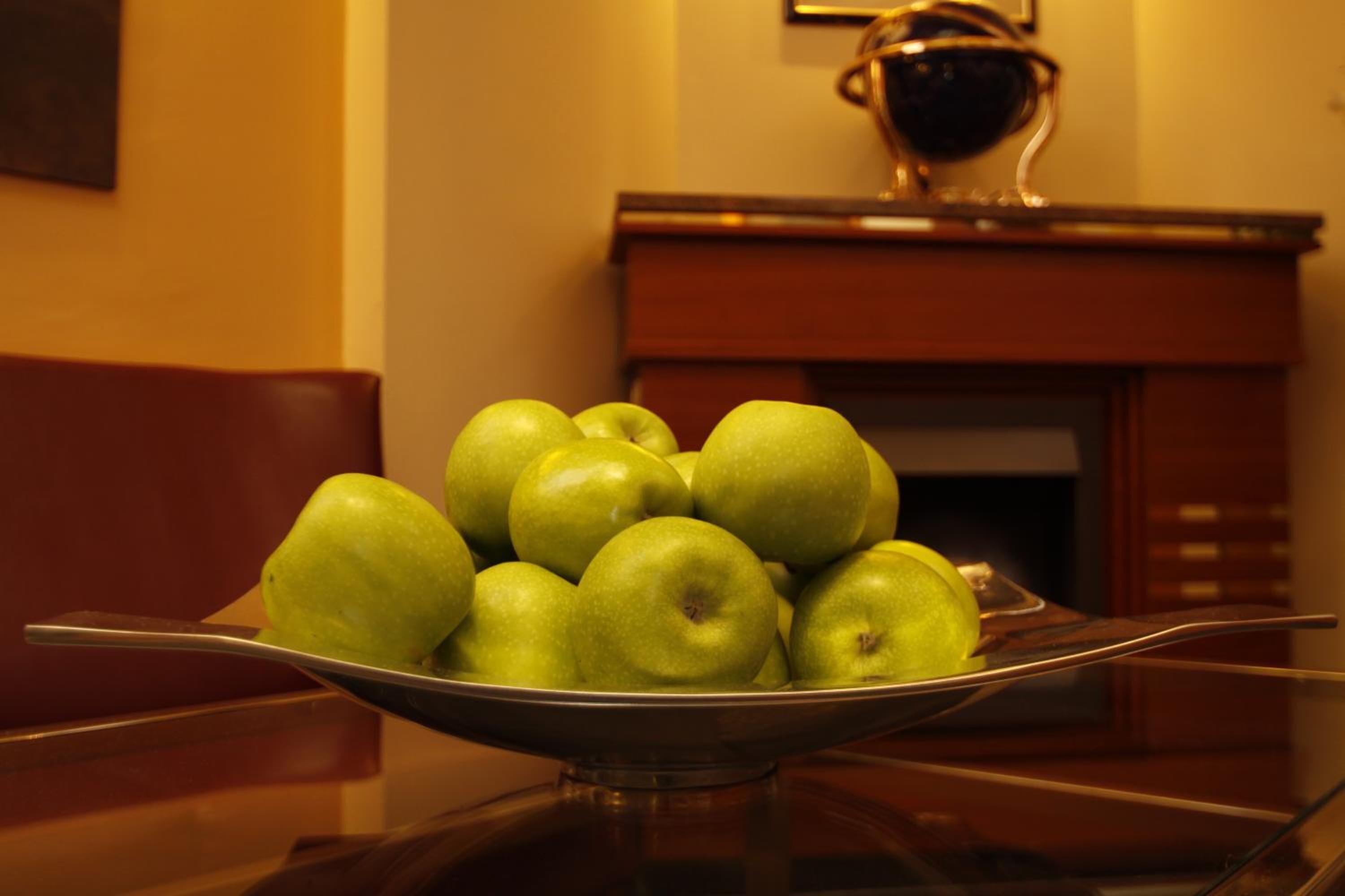 A bowl of green apples on a table with a fireplace in the background.