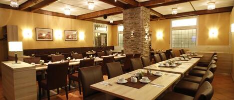A spacious dining area with long tables, wooden beams, stone pillar, and wall-mounted lights.