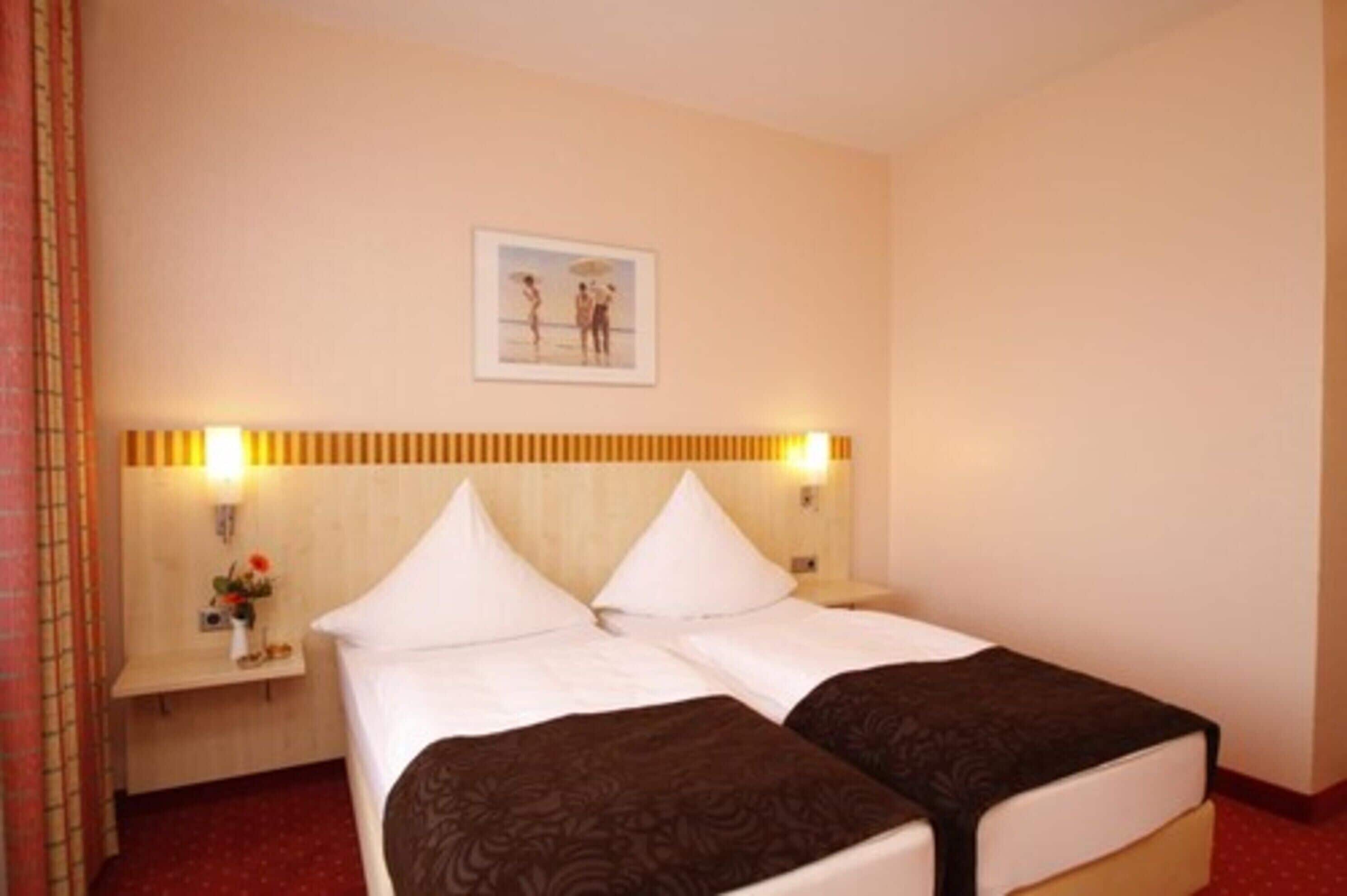 Habitación de hotel con dos camas, una mesita de noche con lámpara, un cuadro en la pared y una pequeña mesa con un florero.